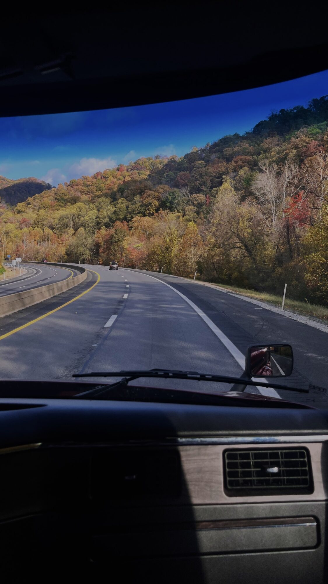 View from inside a vehicle showing a winding road with trees displaying fall colors, and a dashboard with a device attached.