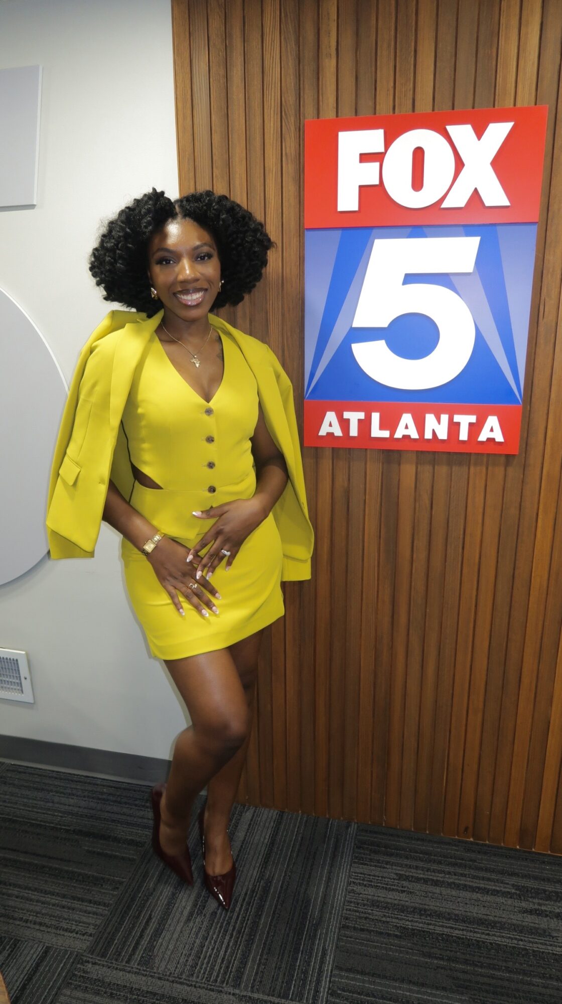 Woman in yellow dress and blazer standing next to FOX 5 Atlanta sign, smiling, in a room with wooden wall paneling.