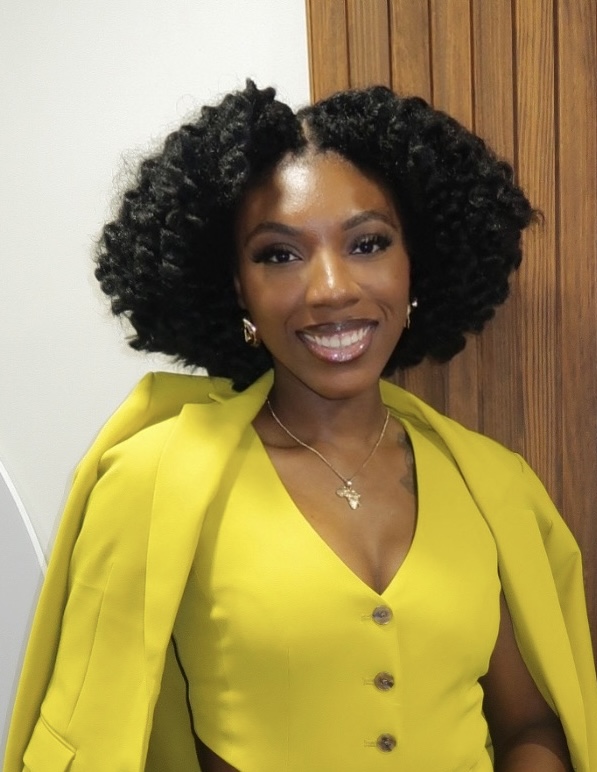 Smiling woman with curly hair wearing a yellow dress and matching blazer, standing indoors near wooden paneling.