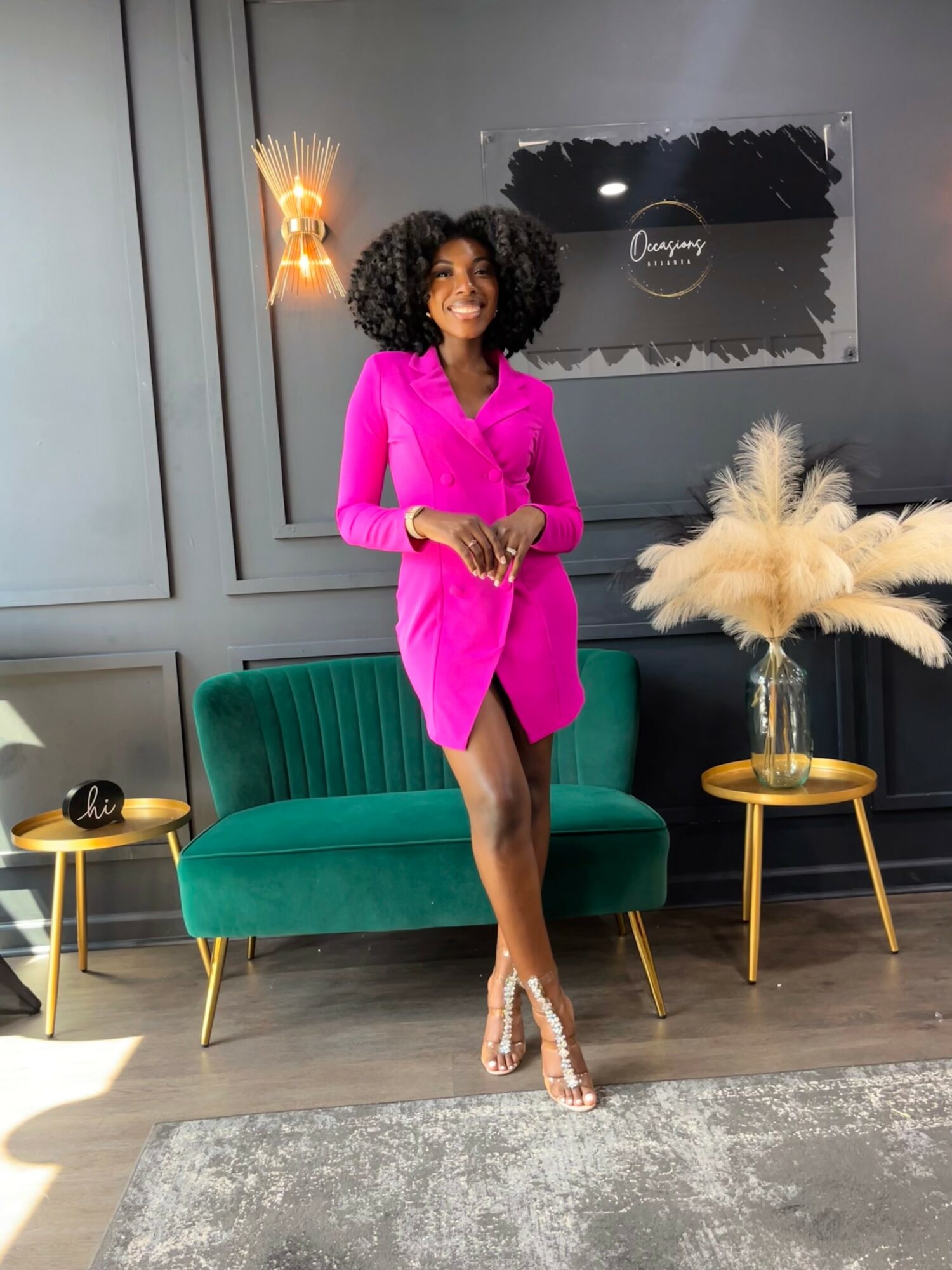 Woman in pink dress standing in stylish room with green sofa, gold side tables, and decorative plants, smiling.