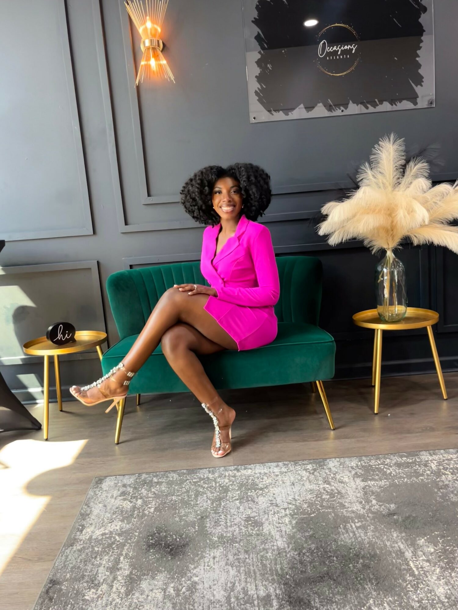 Woman in pink dress sitting on green sofa with gold side tables and decorative plant, gray wall background.