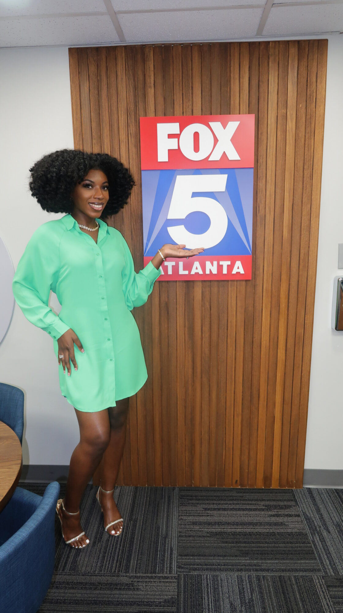 Woman in a green dress standing next to a FOX 5 Atlanta sign on a wooden wall, pointing at it.
