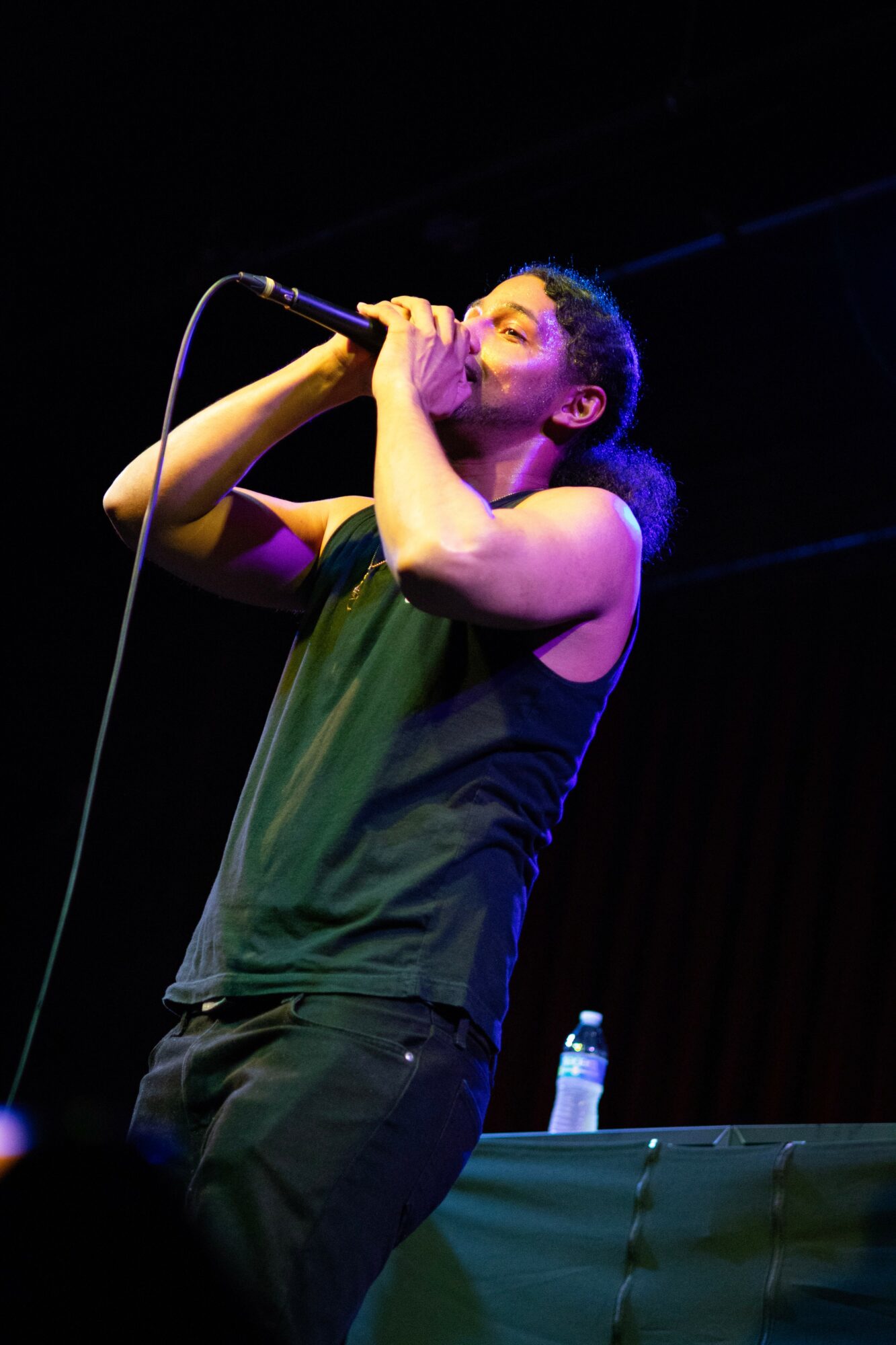 Person singing into a microphone on stage with a water bottle nearby, dark background, stage lighting.