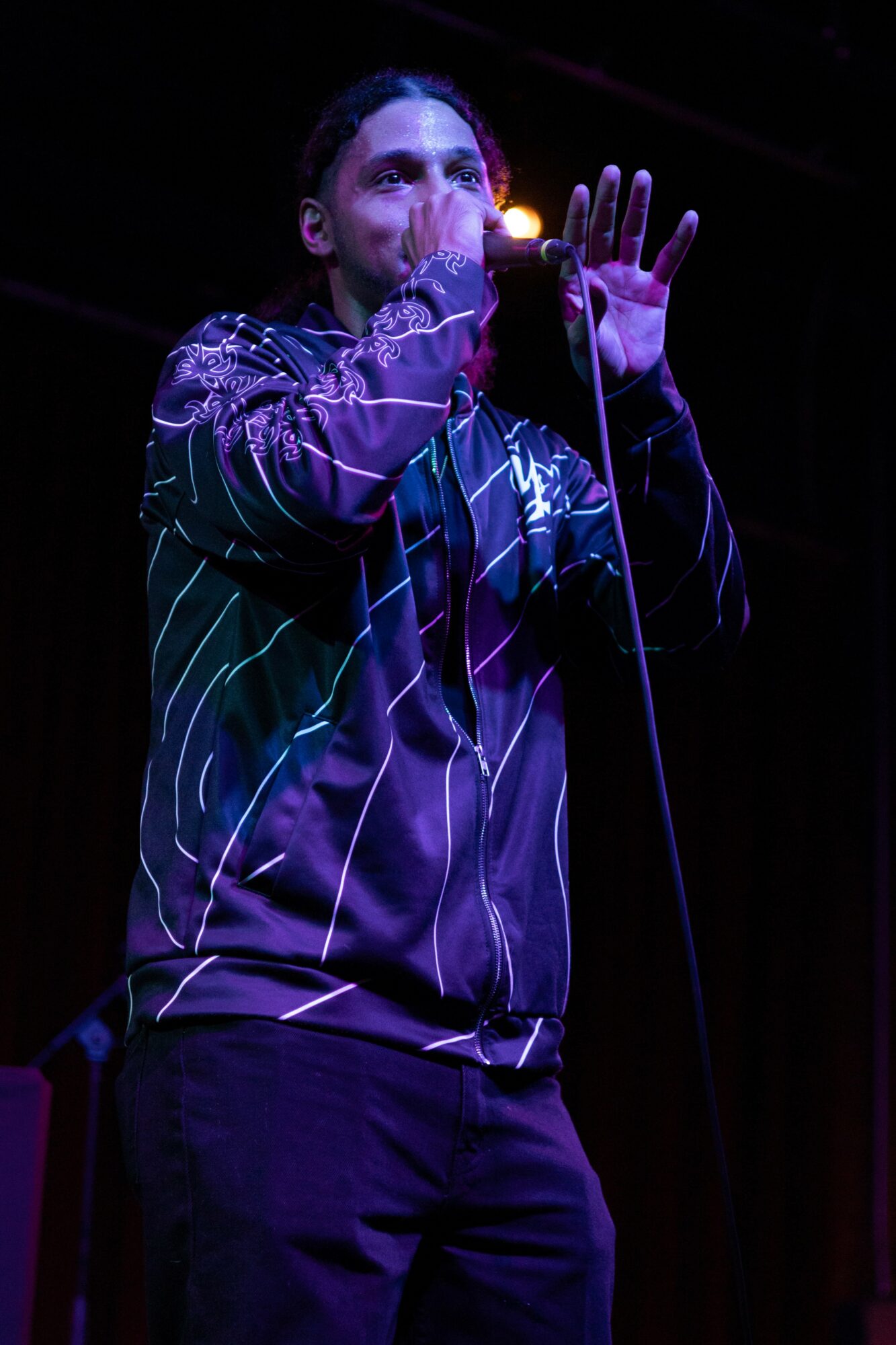 Person performing on stage, holding microphone, wearing a shiny, striped jacket, with dark background.