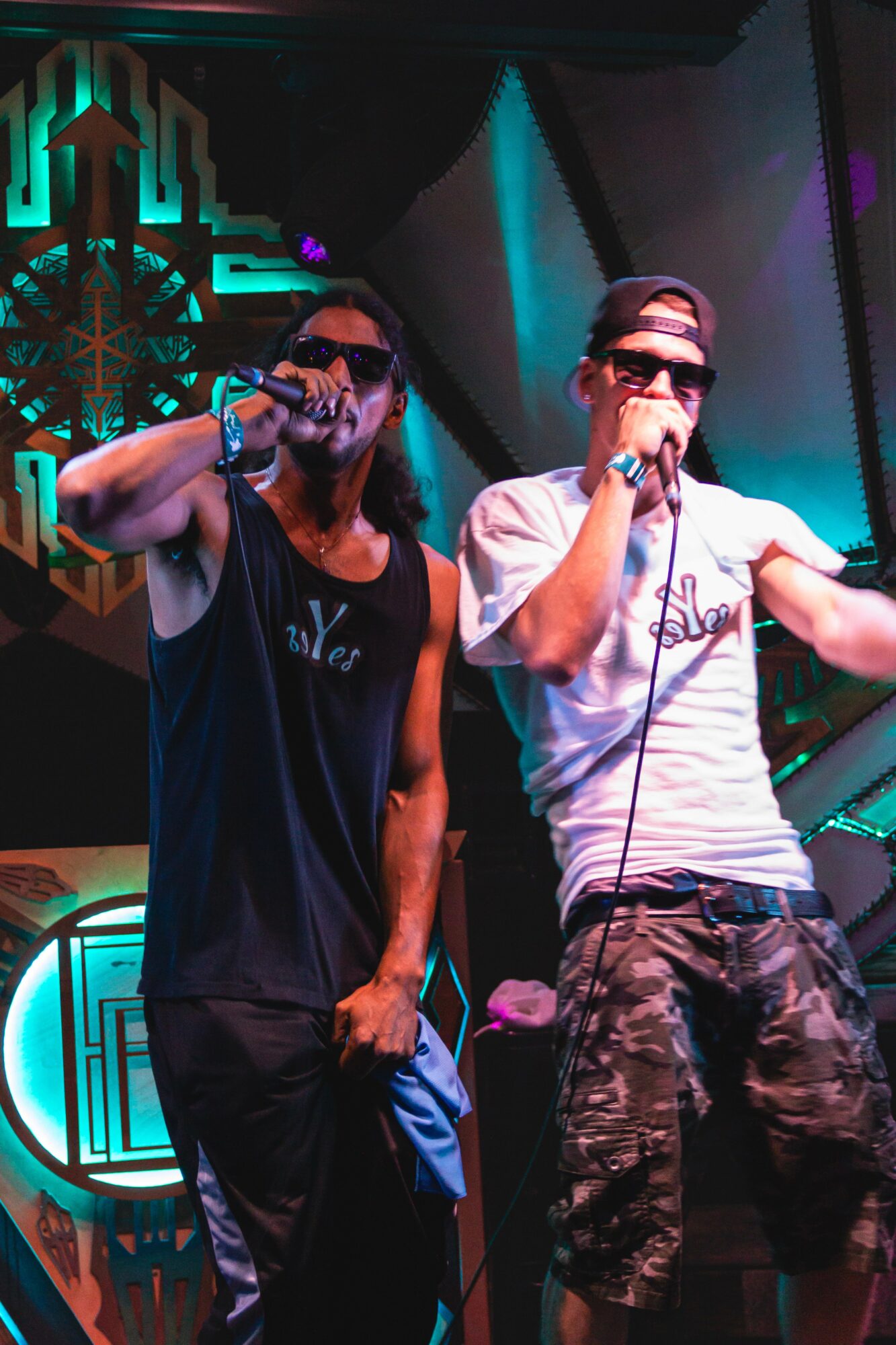 Two men wearing sunglasses and casual clothing perform on stage with microphones, colorful lighting in the background.