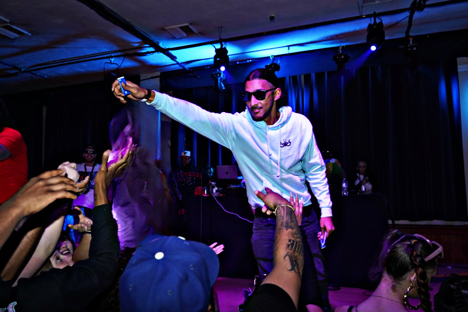 Performer on stage wearing sunglasses and a white hoodie, engaging with audience in a dark, blue-lit setting.