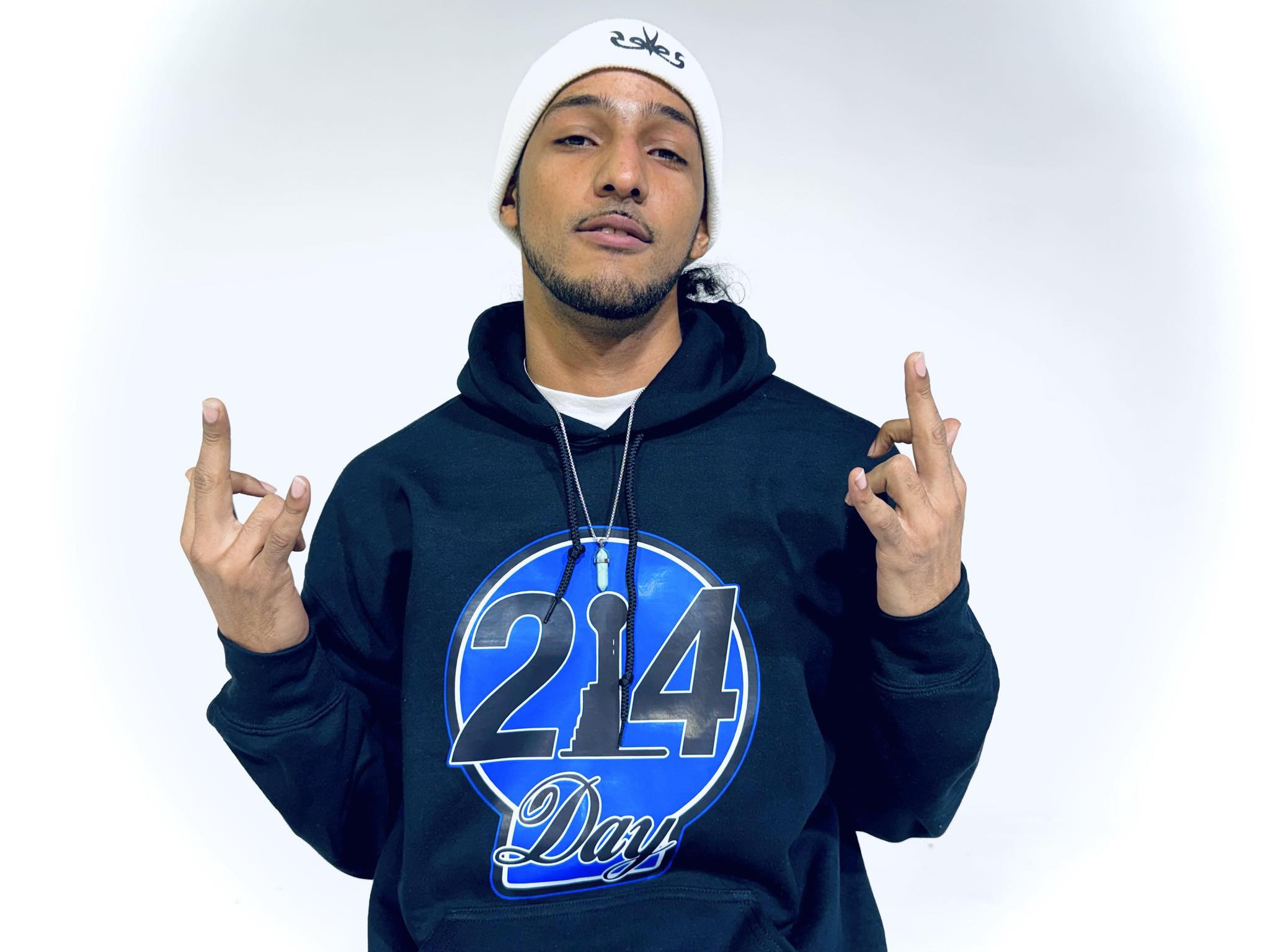 Young man wearing a white beanie and black hoodie with a blue logo, making hand gestures, against a plain background.