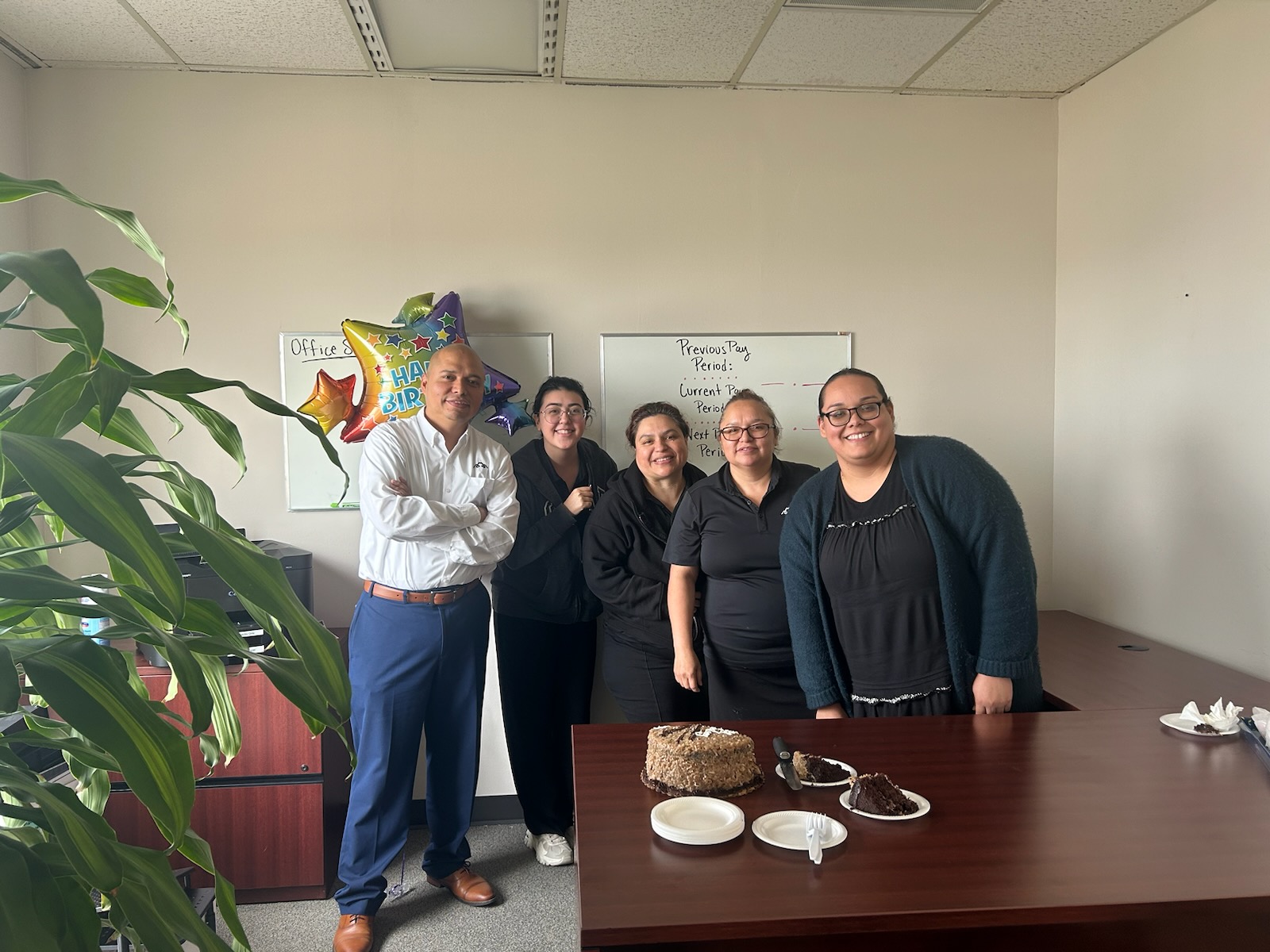 Five people standing together in an office, smiling, with balloons and a whiteboard in the background.