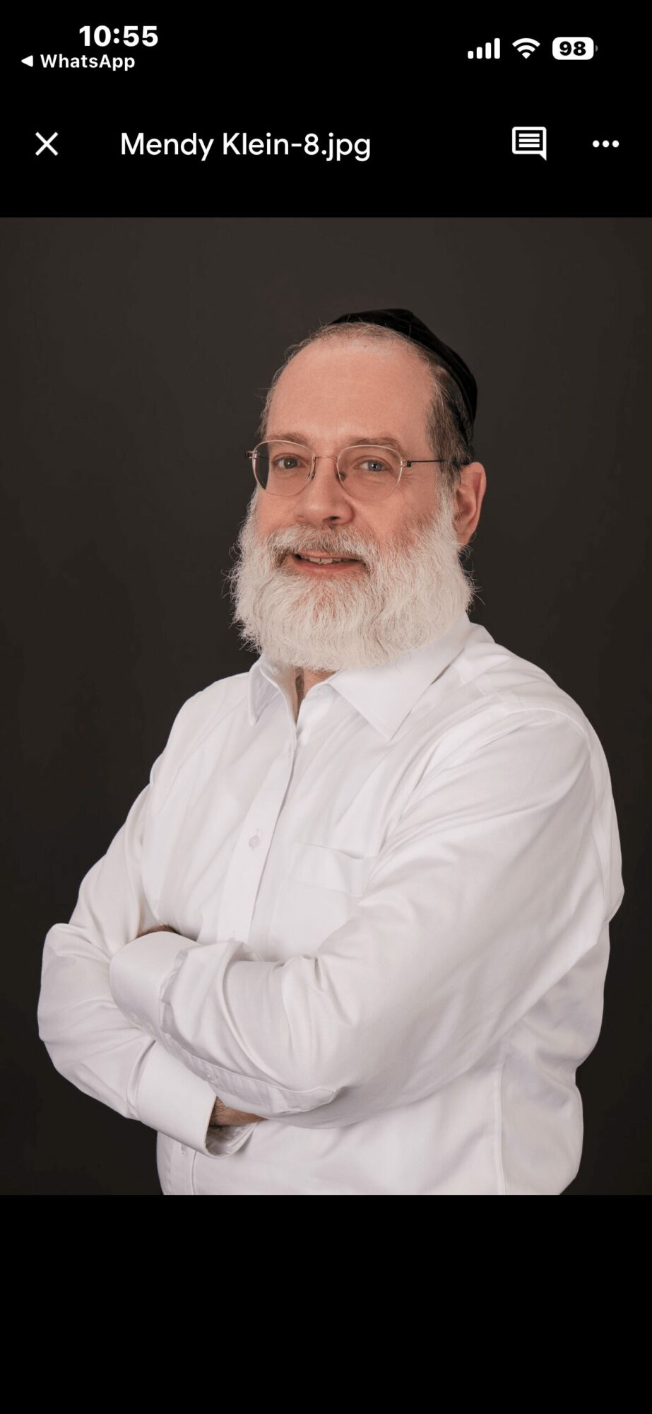 Man with glasses and a beard, wearing a white shirt, standing with arms crossed against a dark background.
