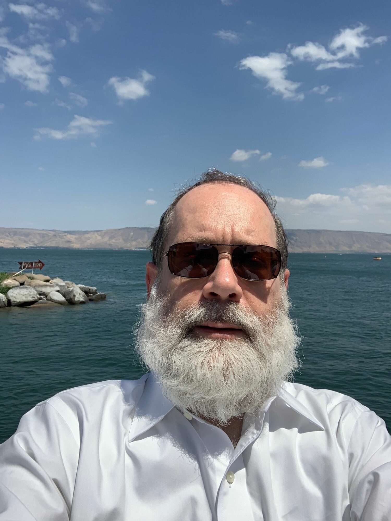 Elderly man with white beard and sunglasses takes a selfie near water, with a boat and shoreline in the background.