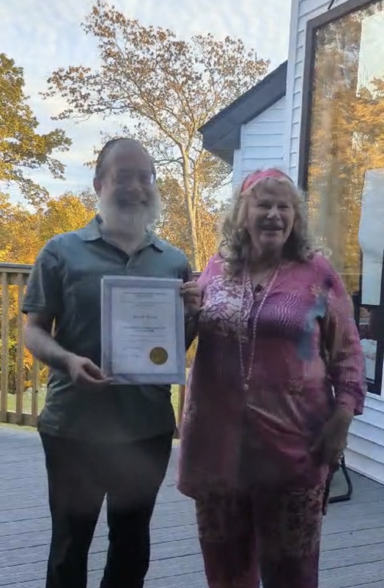 Two women standing outside near a house, one holding a certificate, trees with autumn leaves in background.