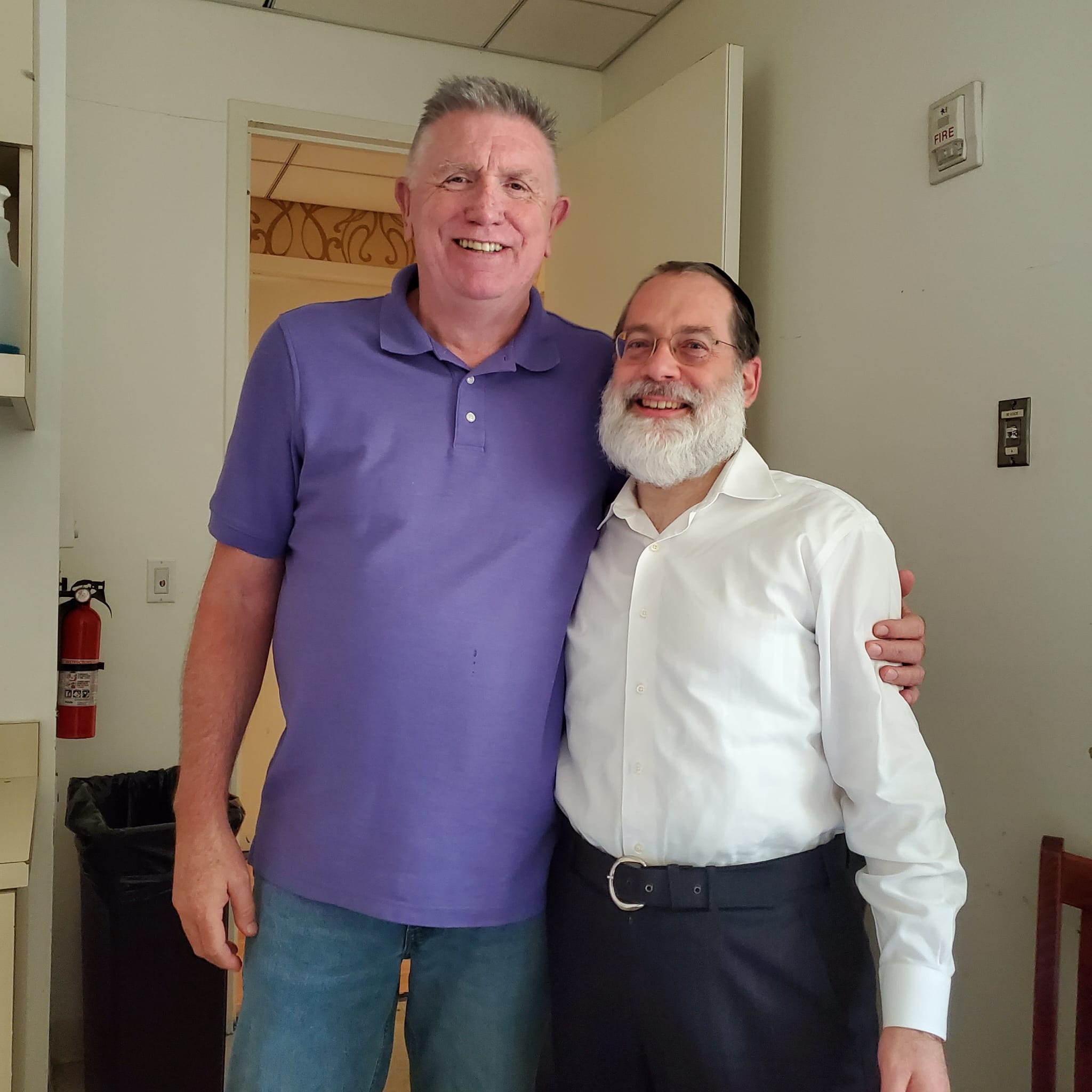 Two men smiling and hugging indoors, one in a purple shirt and jeans, the other in a white shirt and dark pants.