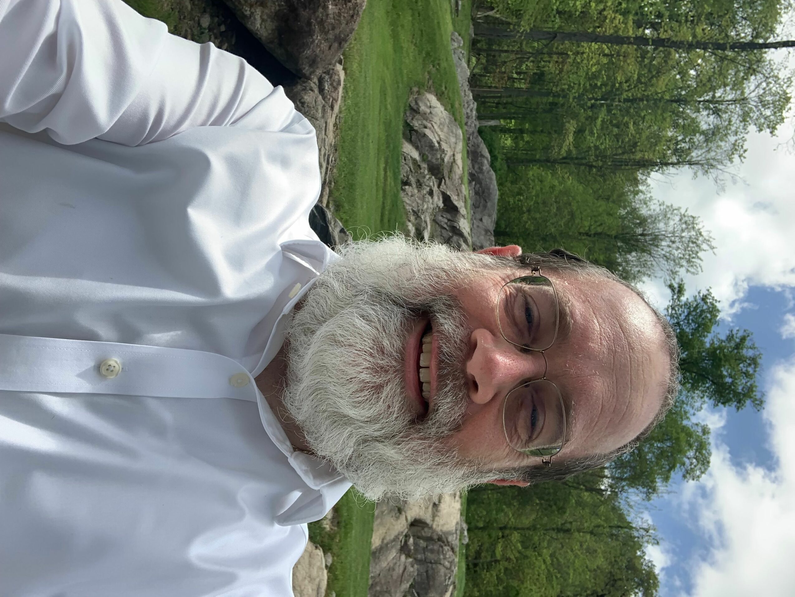 Older man with white beard smiling outdoors, wearing a white shirt, trees and rocks in background.