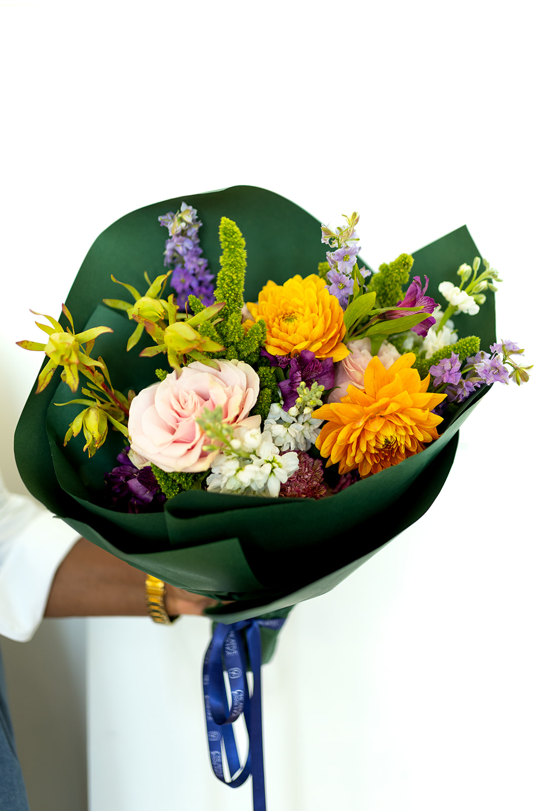 Person holding a colorful bouquet of flowers with yellow, purple, pink, and orange blooms, wrapped in dark green paper.