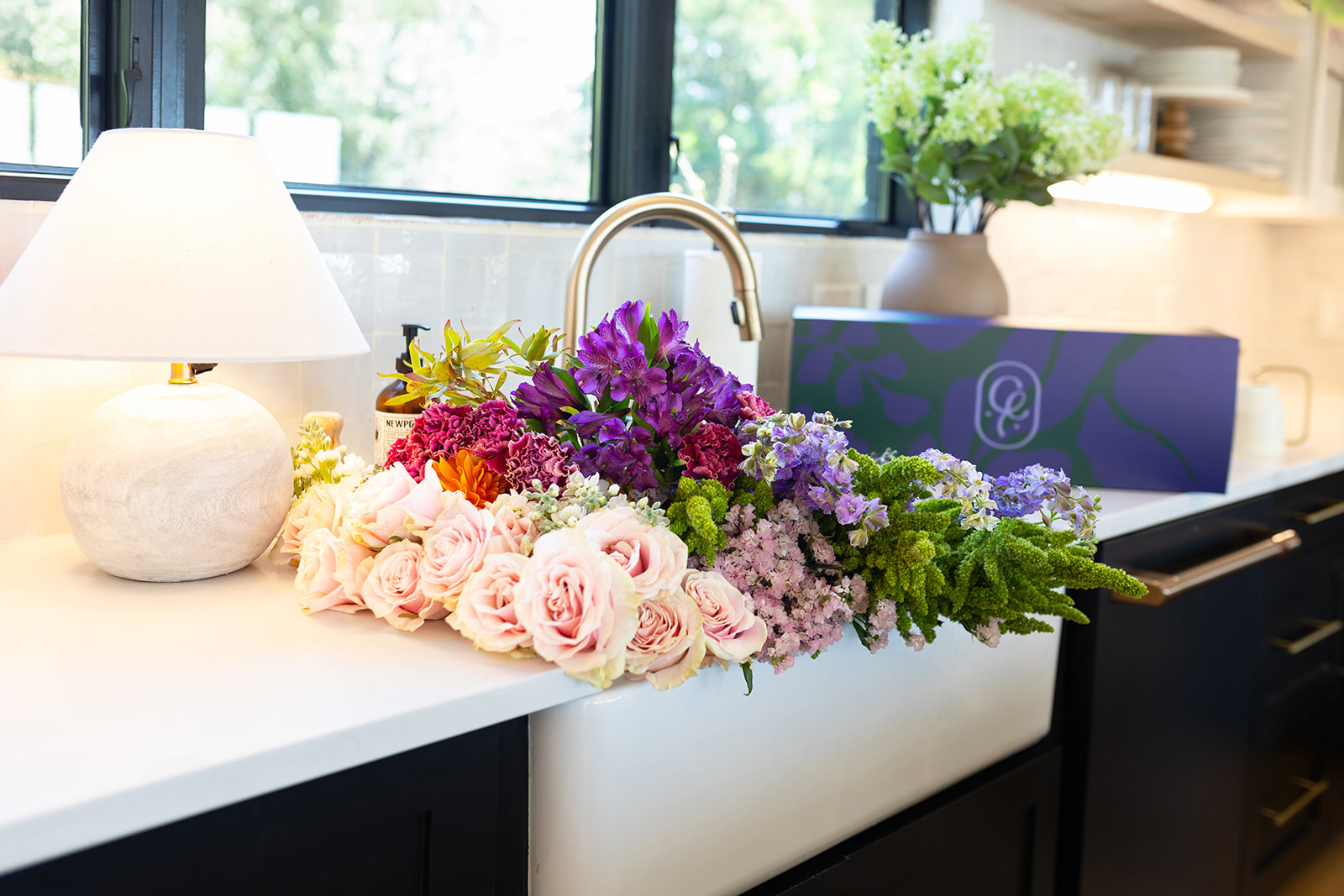 A kitchen countertop with a bouquet of pink, purple, and yellow flowers, a white lamp, and a window with greenery outside.