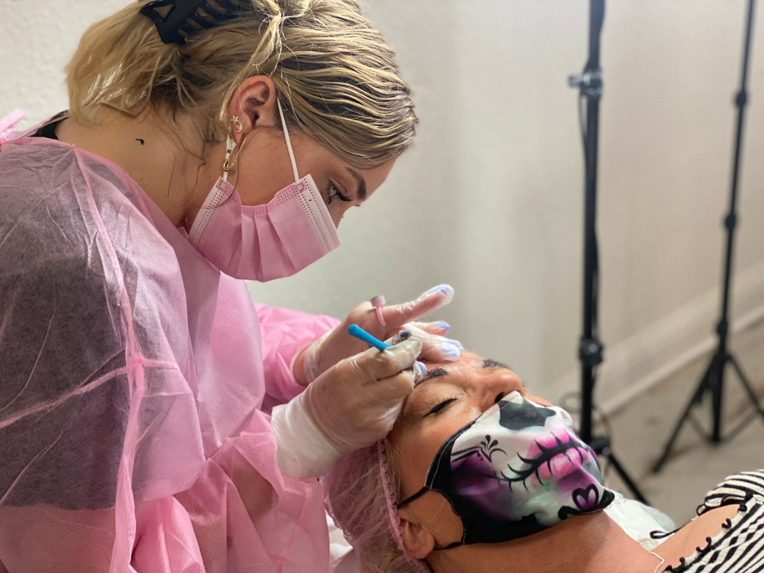 A healthcare worker in pink protective gear and a face mask performs a procedure on a patient wearing a mask, in a clinical setting.