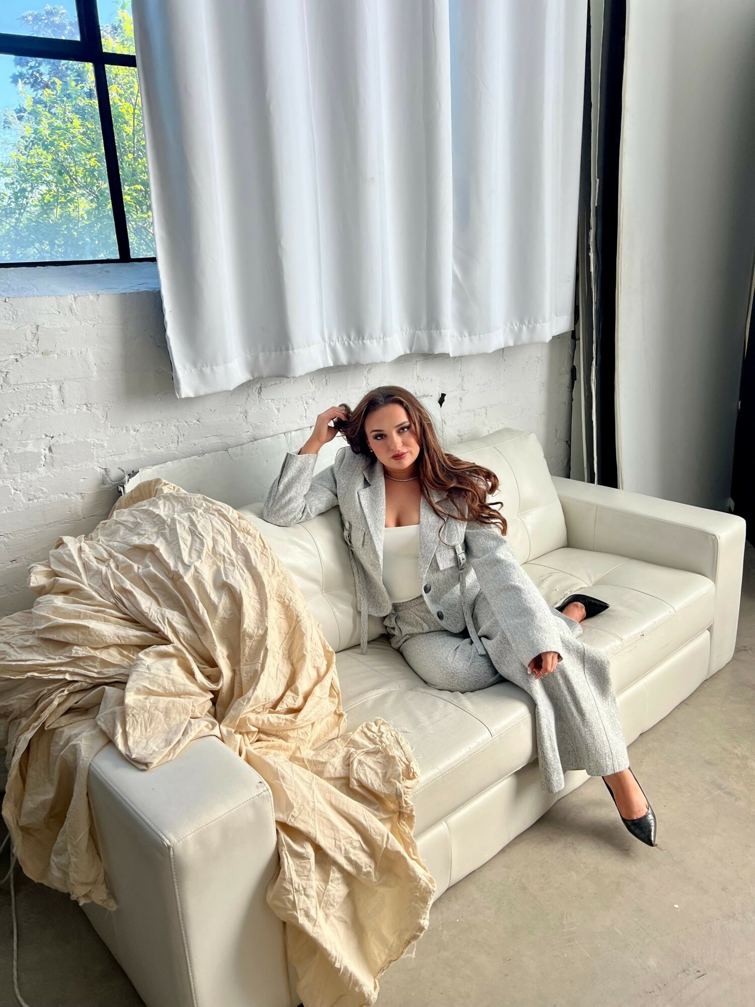 Woman in gray suit sitting on white sofa with beige blanket, in a room with white brick wall and window.
