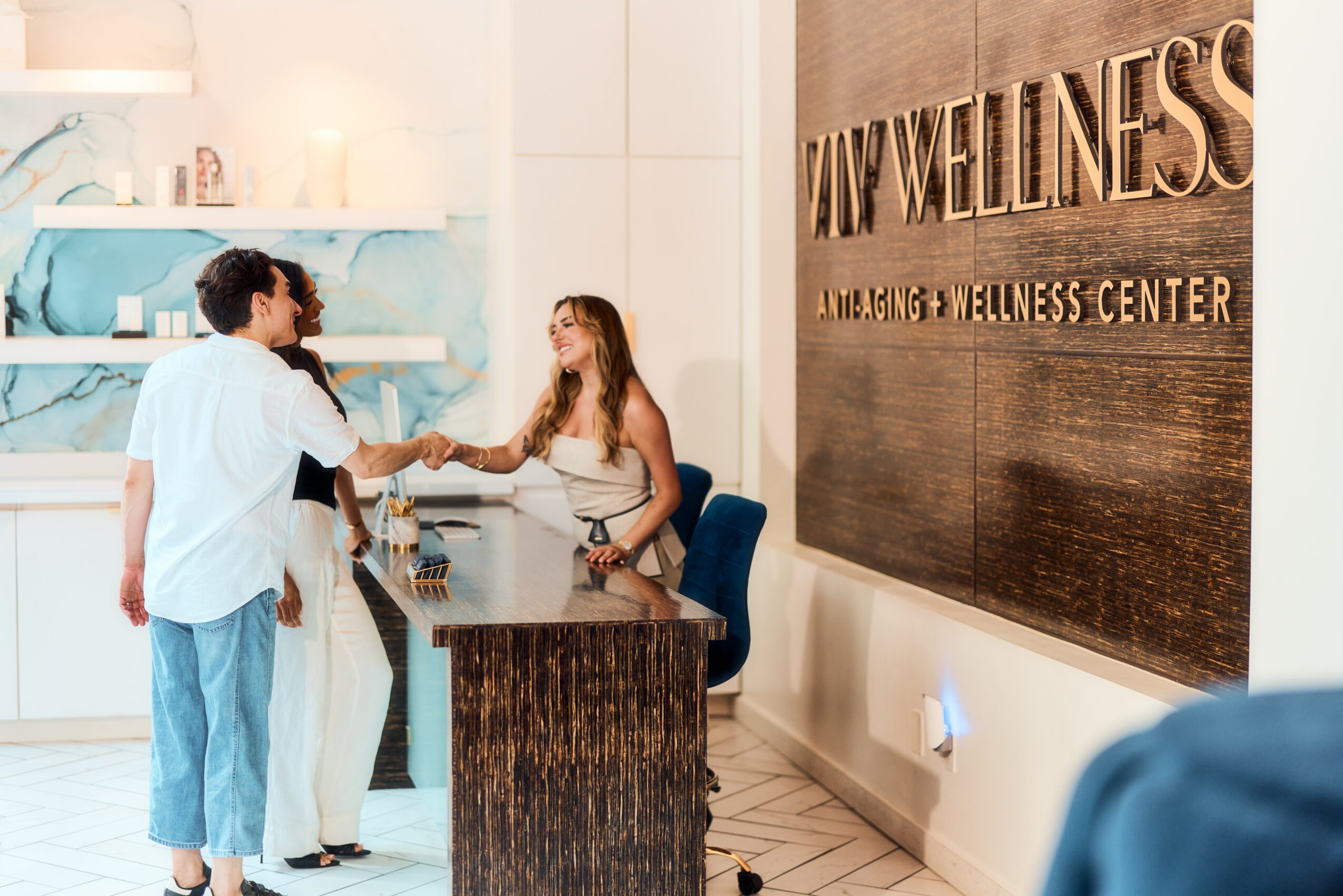 Two women shake hands at a wellness center reception desk, with a large wooden sign reading 'WELLNESS' on the wall.