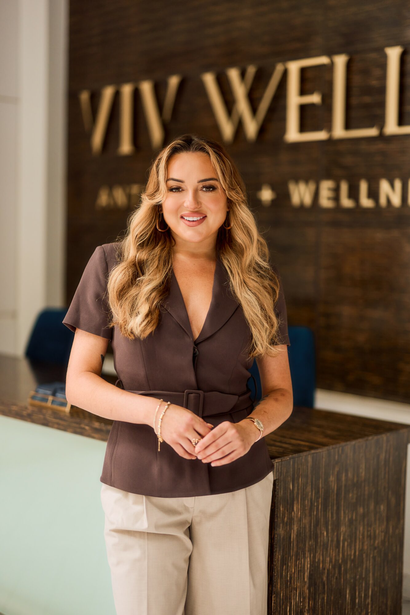Smiling woman with long wavy hair standing in front of a wooden wall with 'VIV WELL' sign, wearing a dark top and light pants.