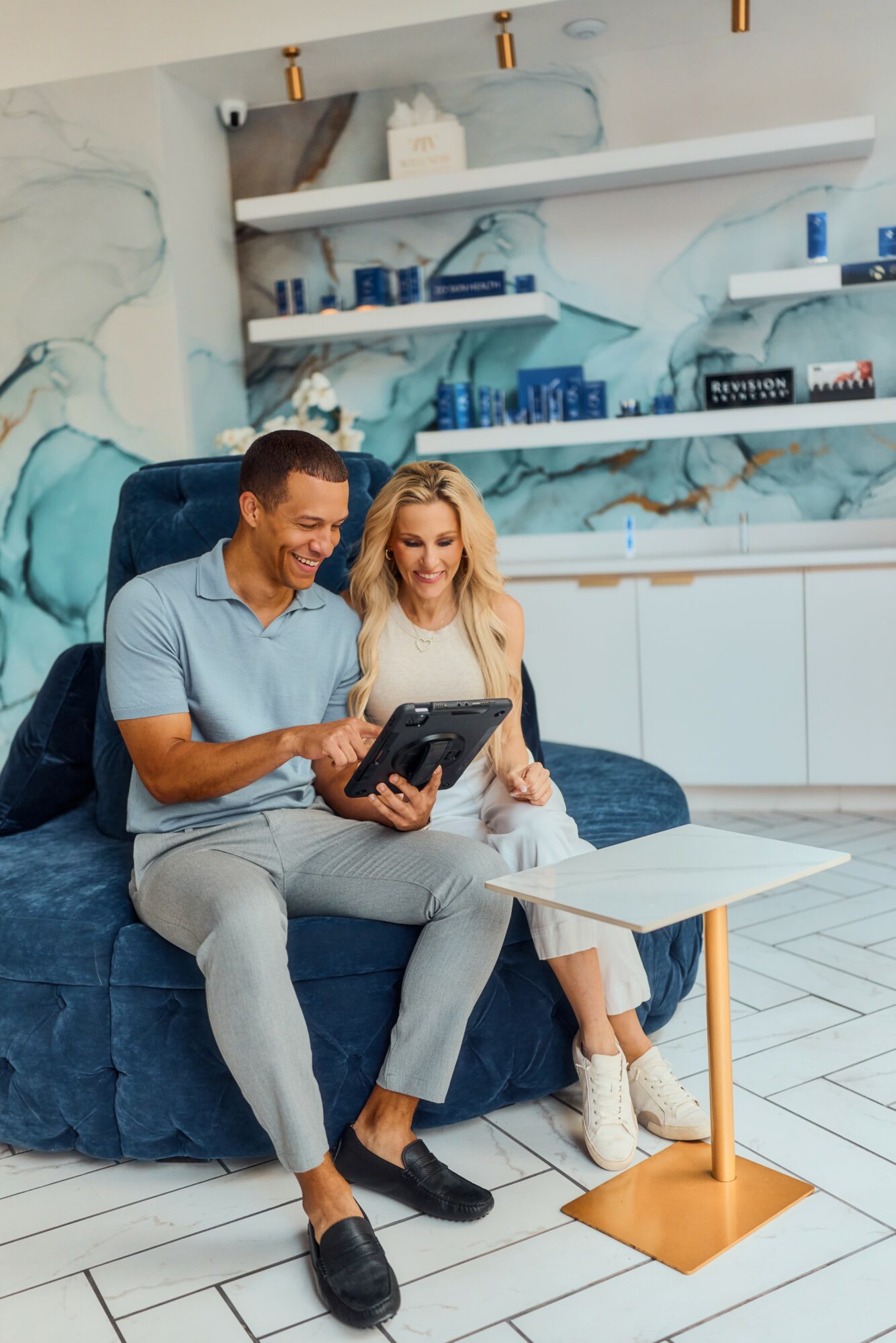 Two people sitting on a blue sofa, looking at a tablet, in a room with a white tiled floor and abstract wall art.