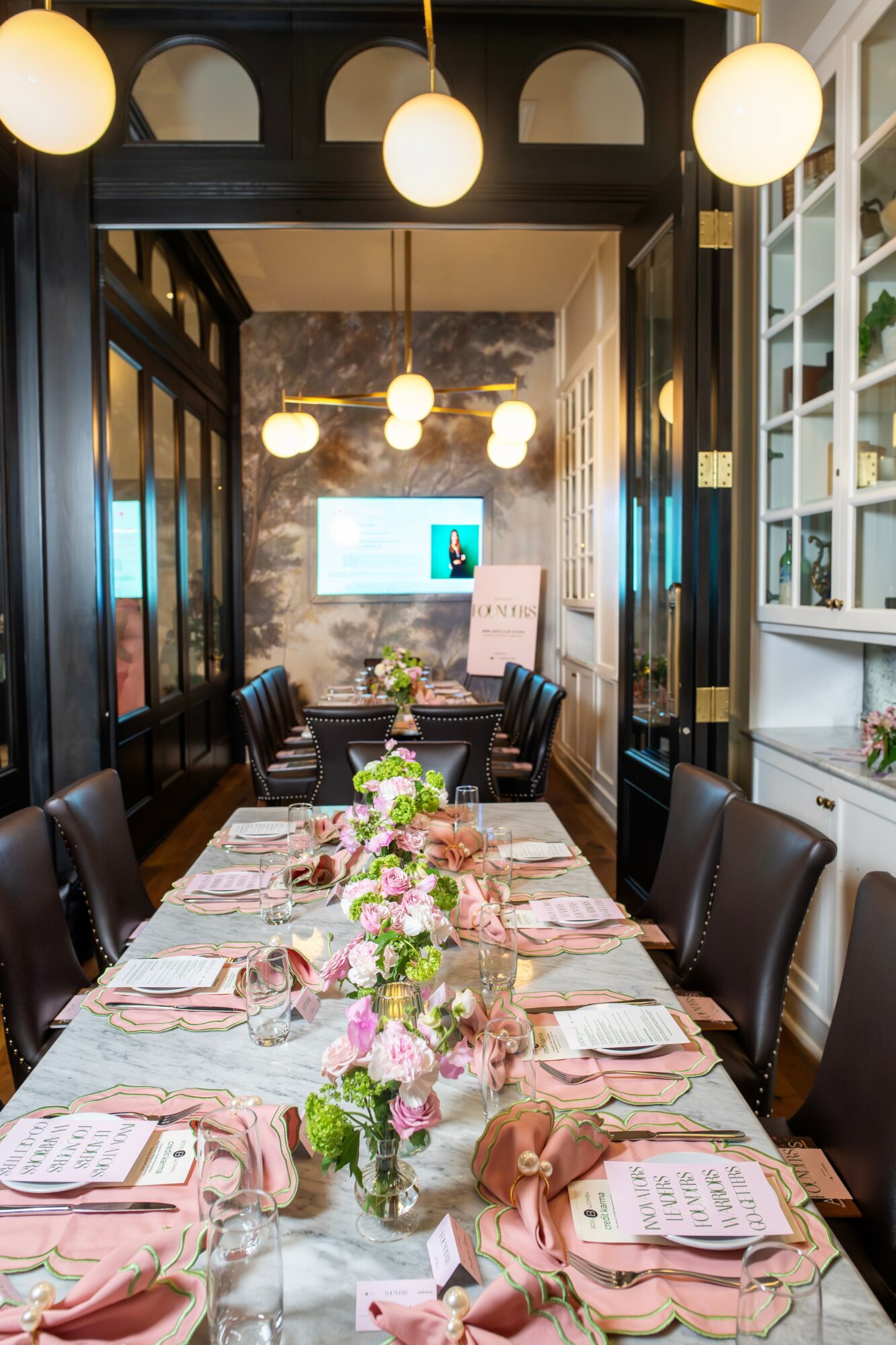 Long dining table set with pink napkins, floral centerpieces, and place settings in a decorated room with hanging lights and a TV screen.