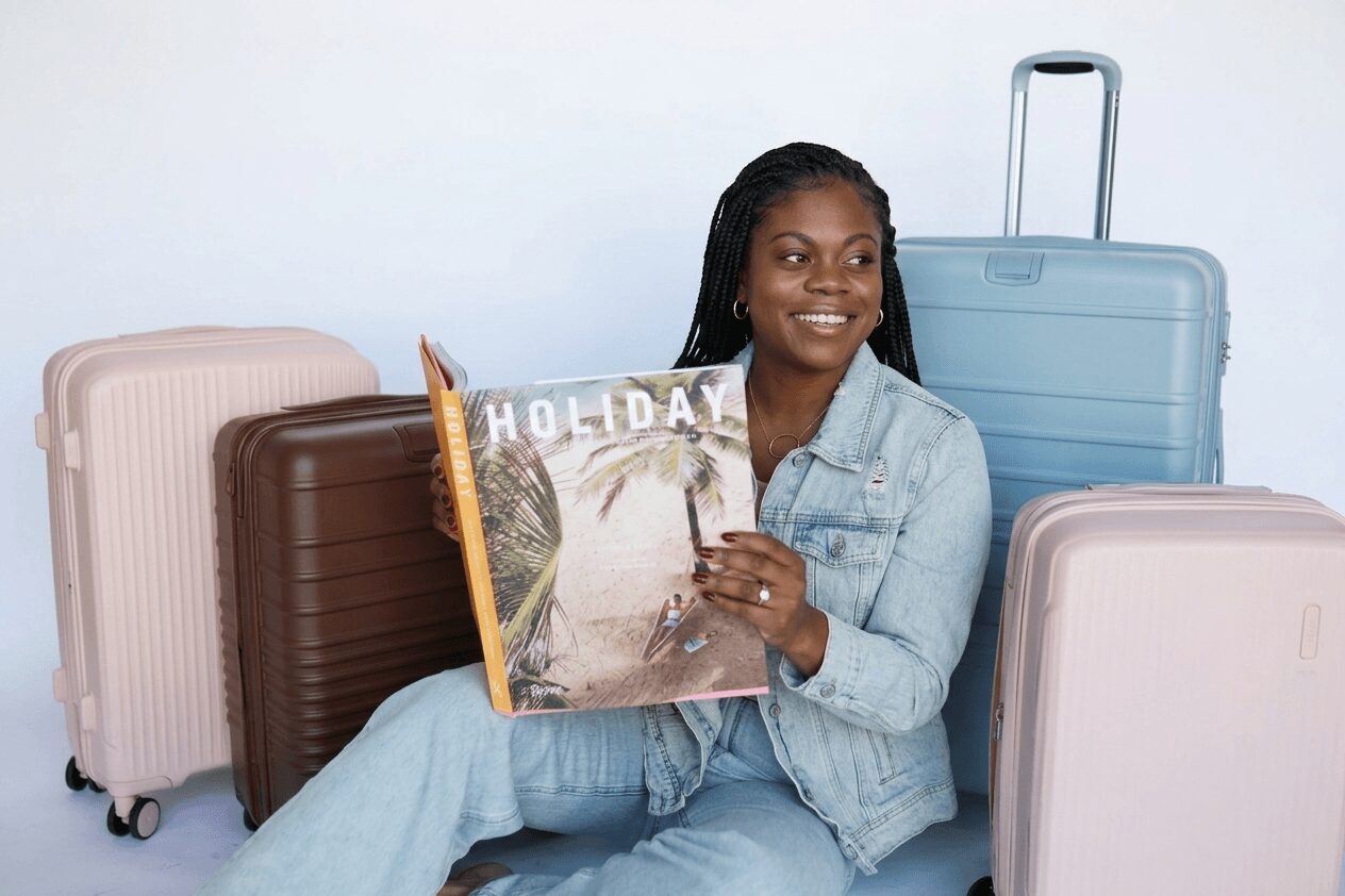 Woman smiling holding a magazine with luggage around her, against a plain background.