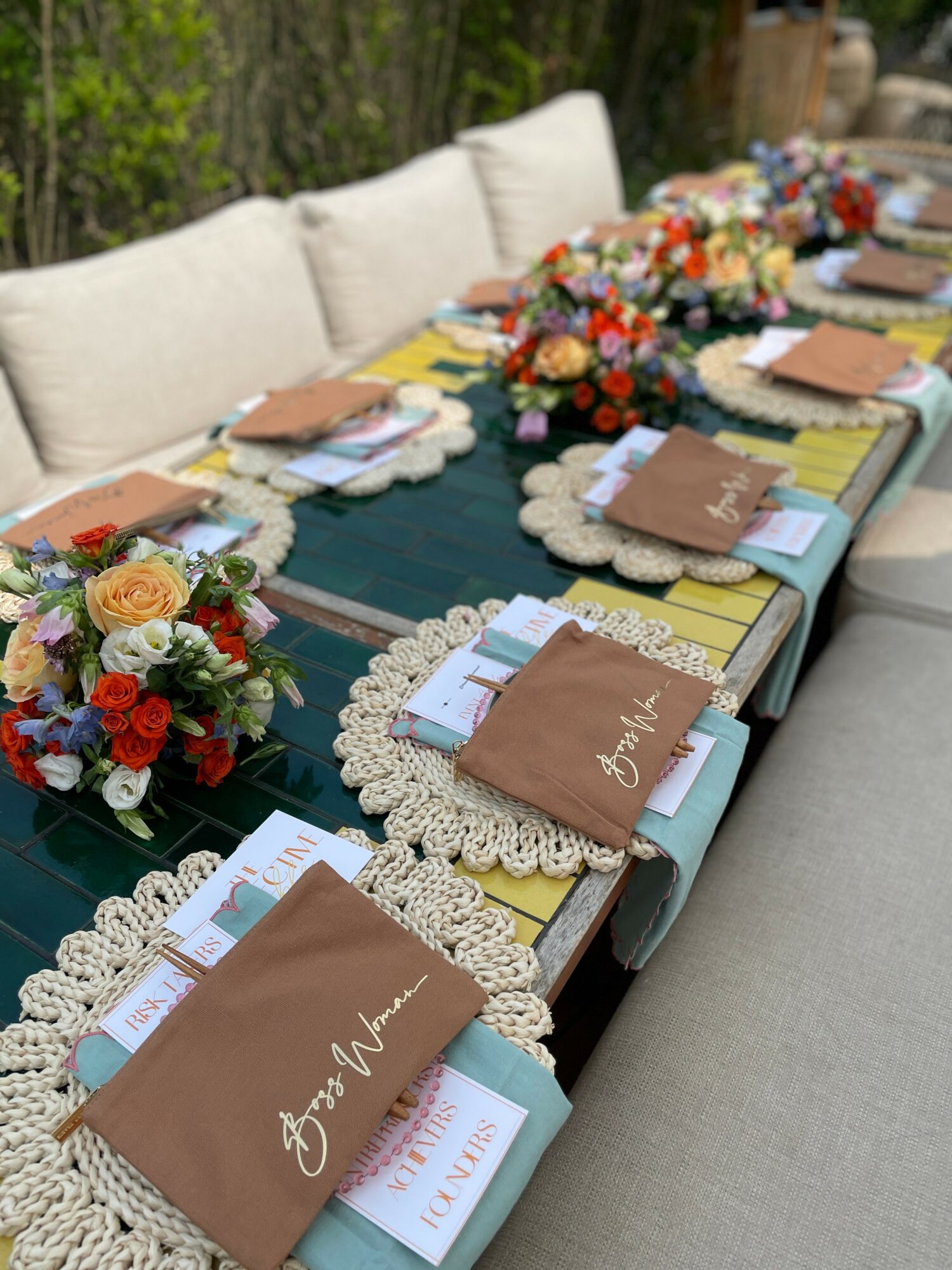 Long table set with floral arrangements, place settings, and decorative doilies, outdoors with a beige sofa in the background.