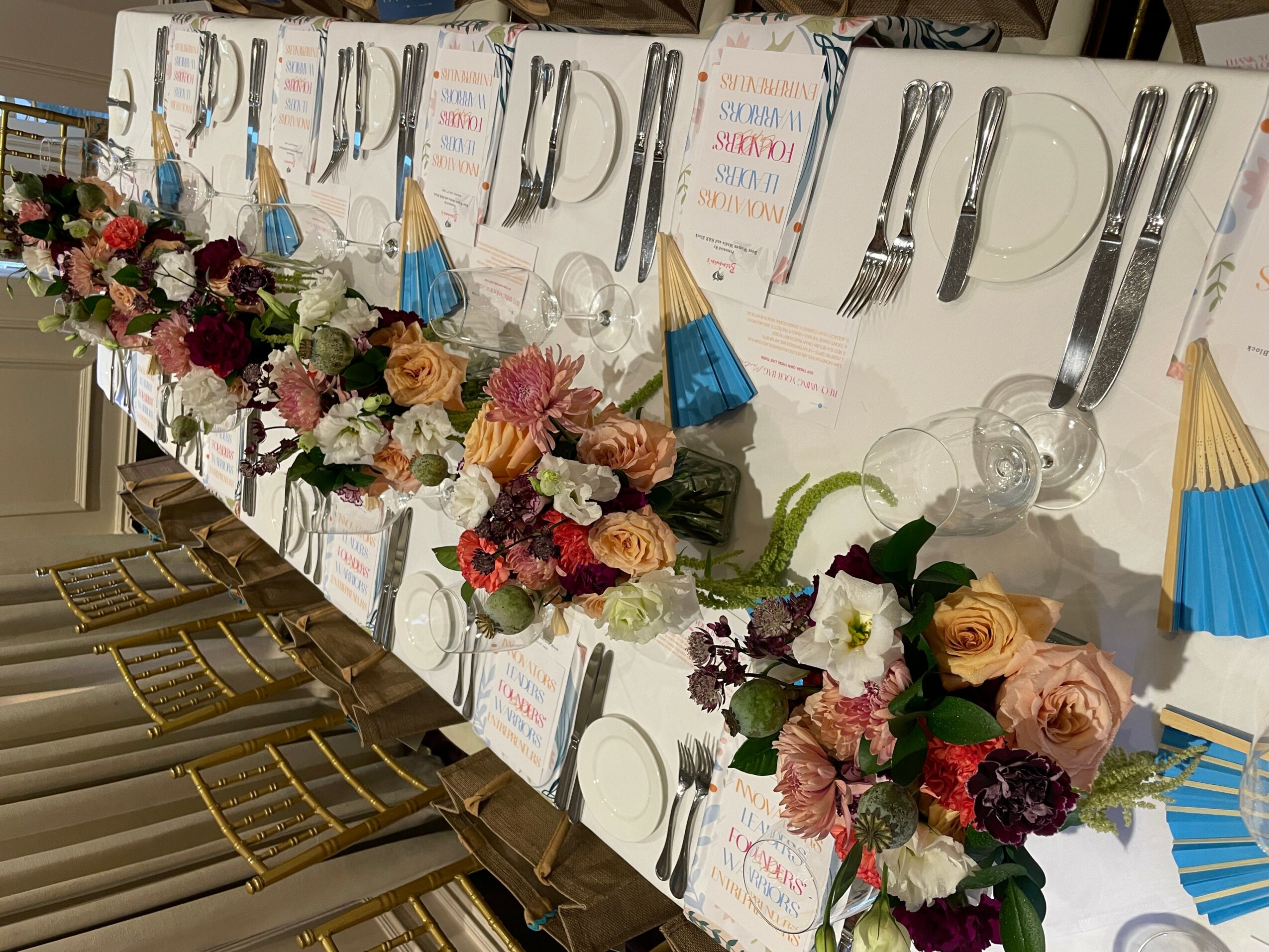 Table decorated with colorful flowers, place cards, and cutlery arranged neatly, with chairs on one side, in a festive setting.