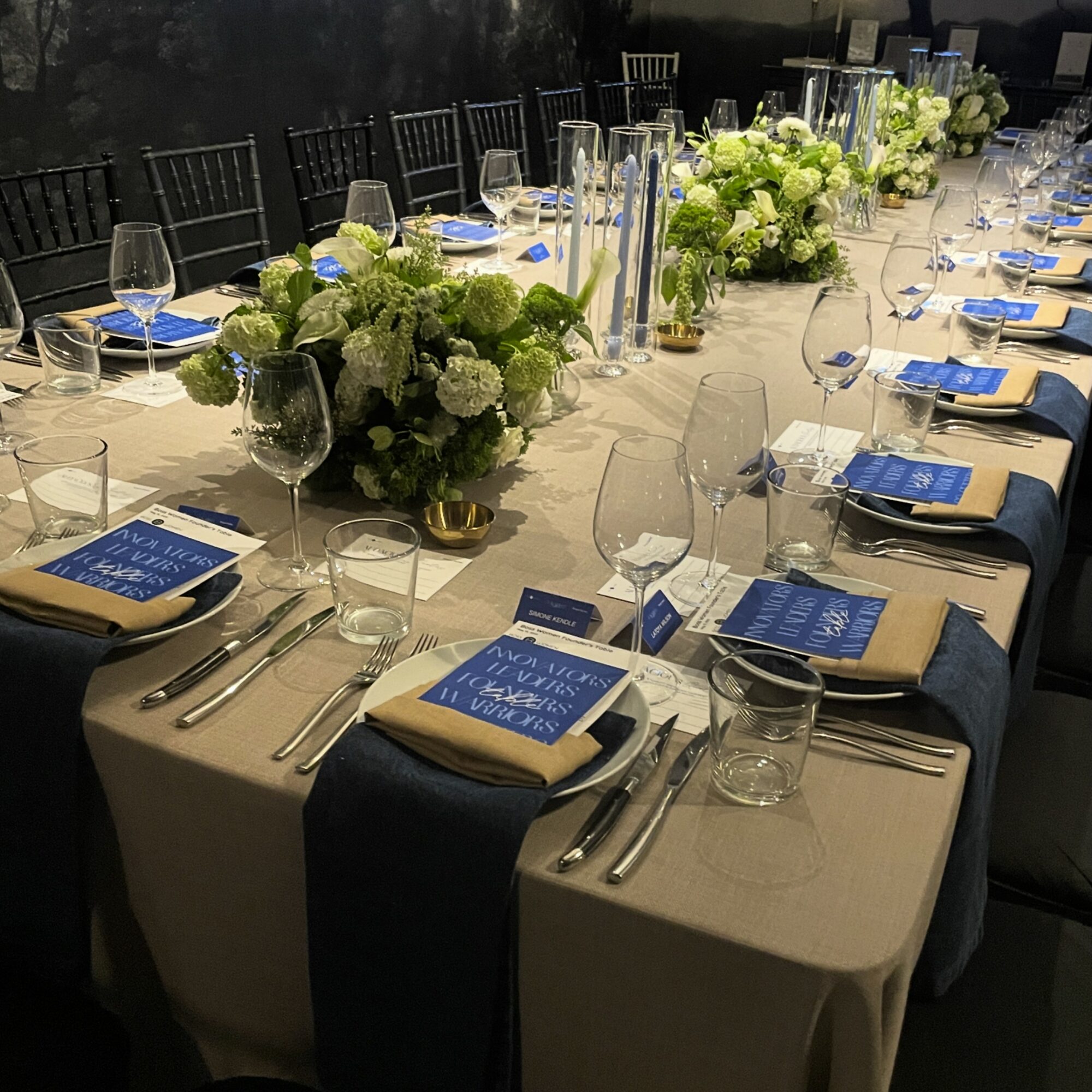Long banquet table set with floral centerpieces, blue menus, wine glasses, and silverware, with black chairs in the background.
