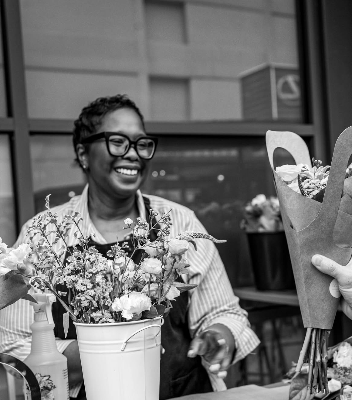 Smiling woman with glasses holding a bouquet of flowers, sitting indoors near a window, with more flowers nearby.
