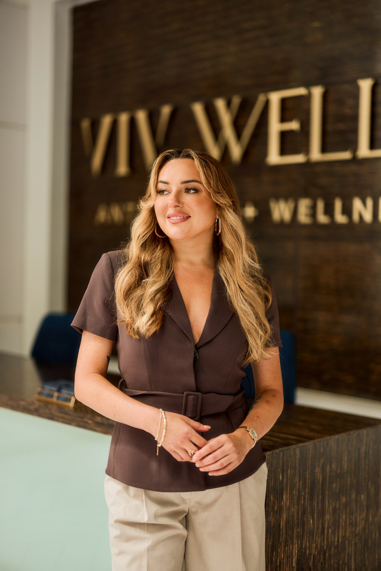 Woman with long wavy hair smiling, standing in front of a wellness center sign, wearing a dark top and light pants.