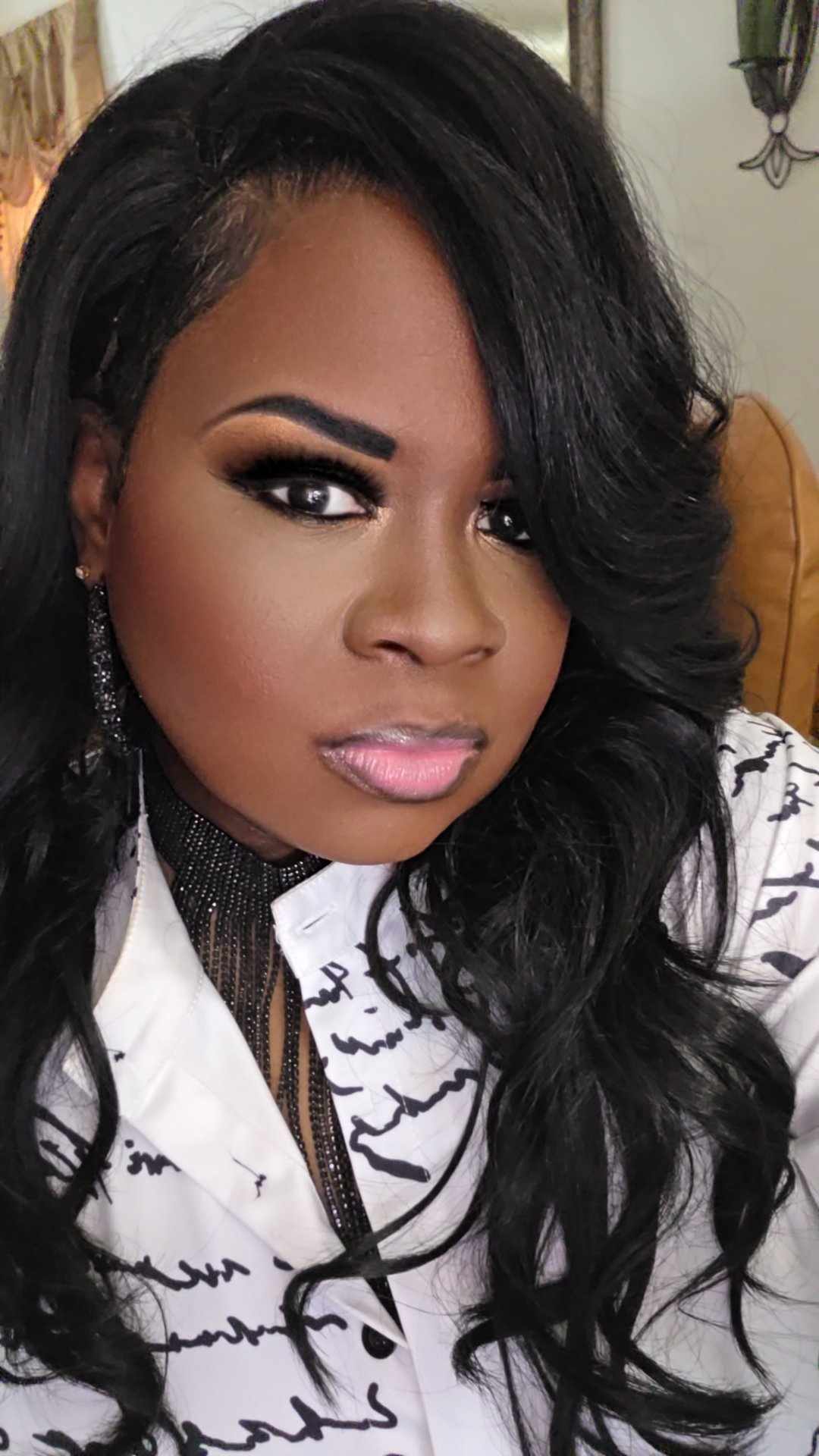 Woman with long black hair, makeup, and earrings, wearing a white patterned top, looking at the camera.