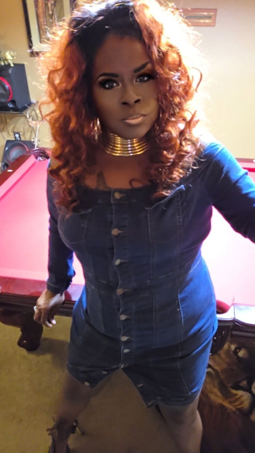 Woman with curly hair wearing a denim dress and gold choker, standing indoors with a pool table in background.