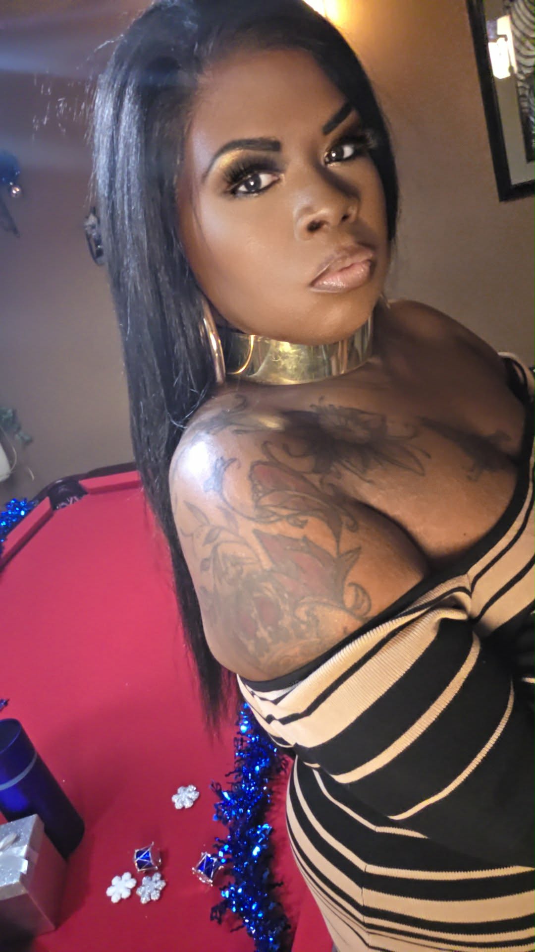 Woman with long dark hair, gold choker, and tattoos on her shoulder and arm, wearing a striped top.