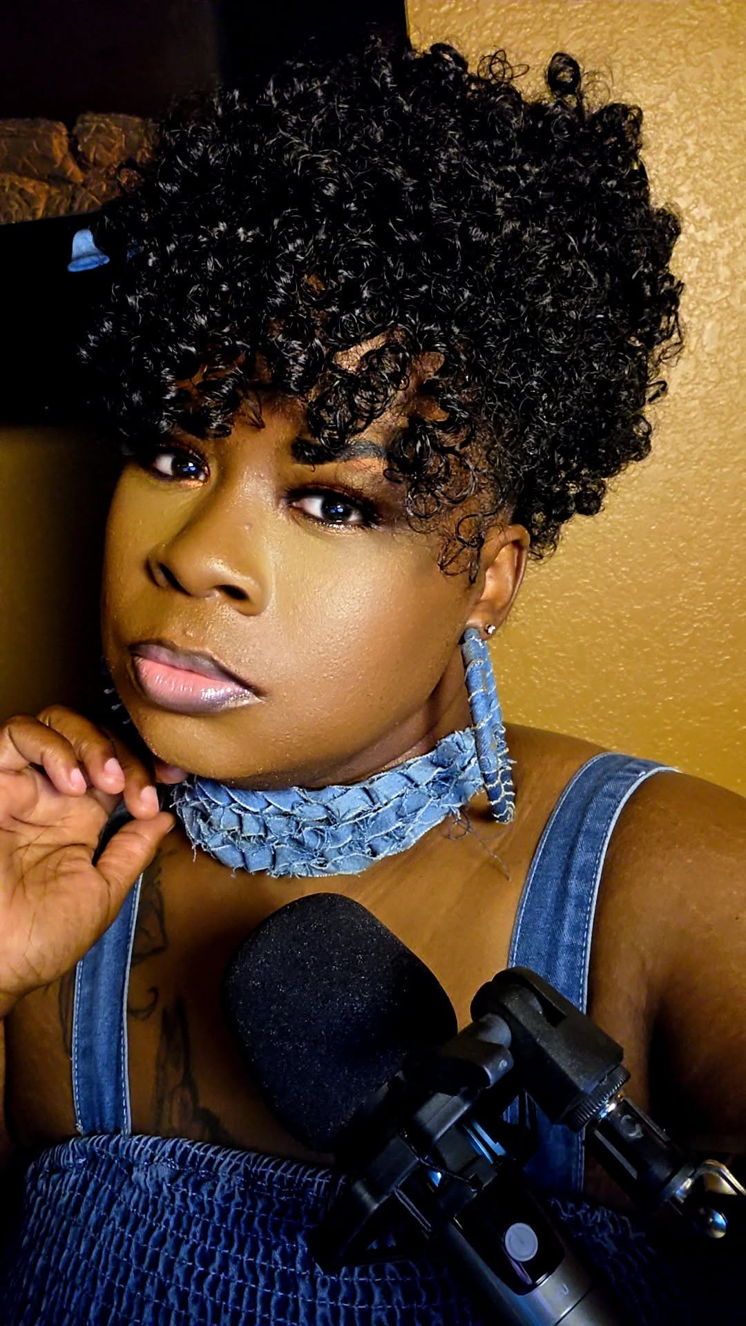 Woman with curly hair, wearing a blue choker and top, holding a microphone, looking at the camera.