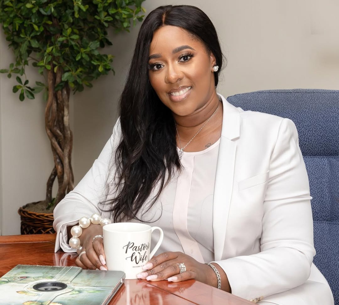 Woman with long dark hair in white blazer sitting at desk with a mug, smiling, with a plant in the background.