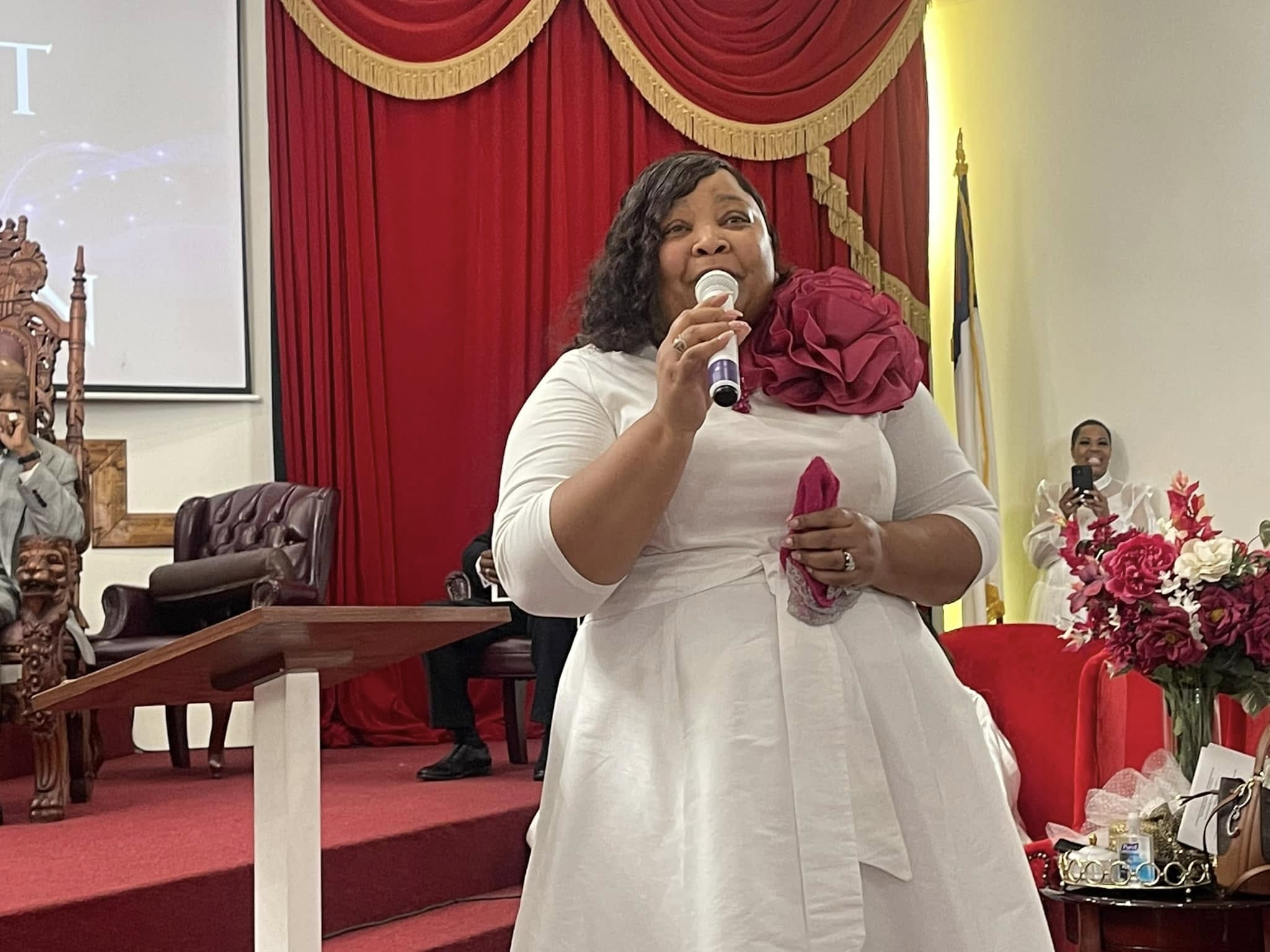 Woman in white dress speaking into microphone, red curtains and floral arrangements in background.