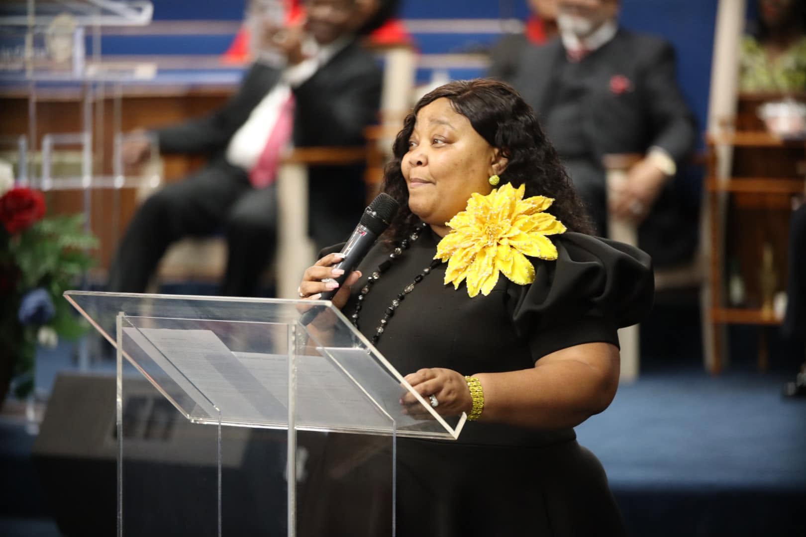 Woman speaking at a podium with a yellow flower on her shoulder, holding a microphone, in a formal setting.
