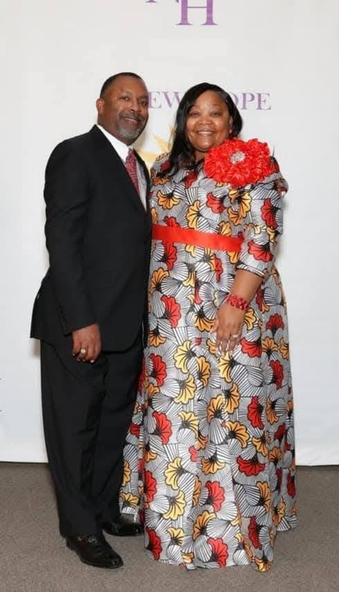 A man and woman stand together, smiling, in front of a white backdrop with purple text, dressed formally.