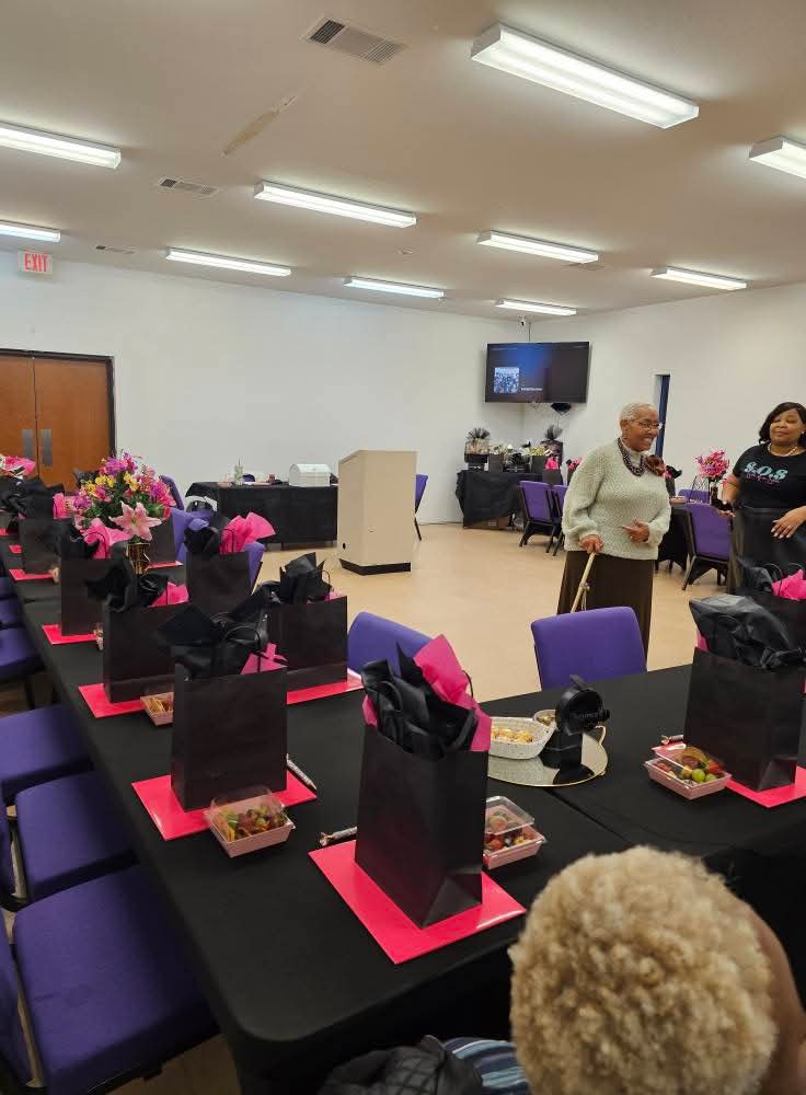 Room with tables set for a celebration, gift bags, and people standing and talking, with a TV on the wall.