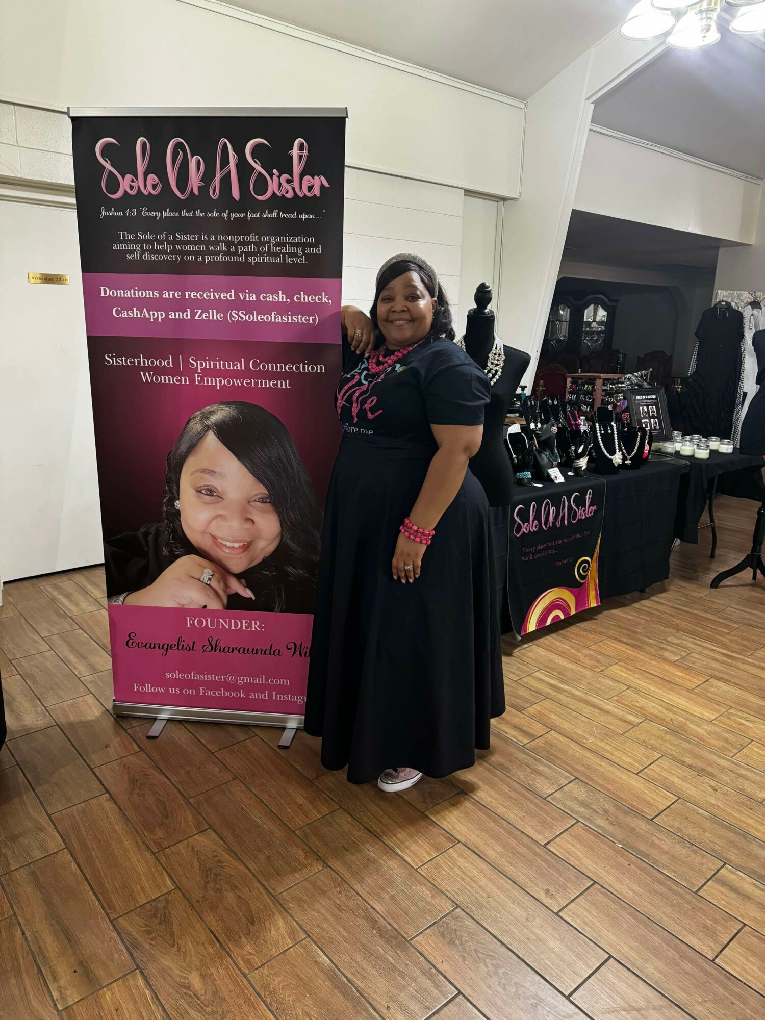 Woman standing next to a pink and black banner at an indoor event, with tables displaying jewelry in the background.