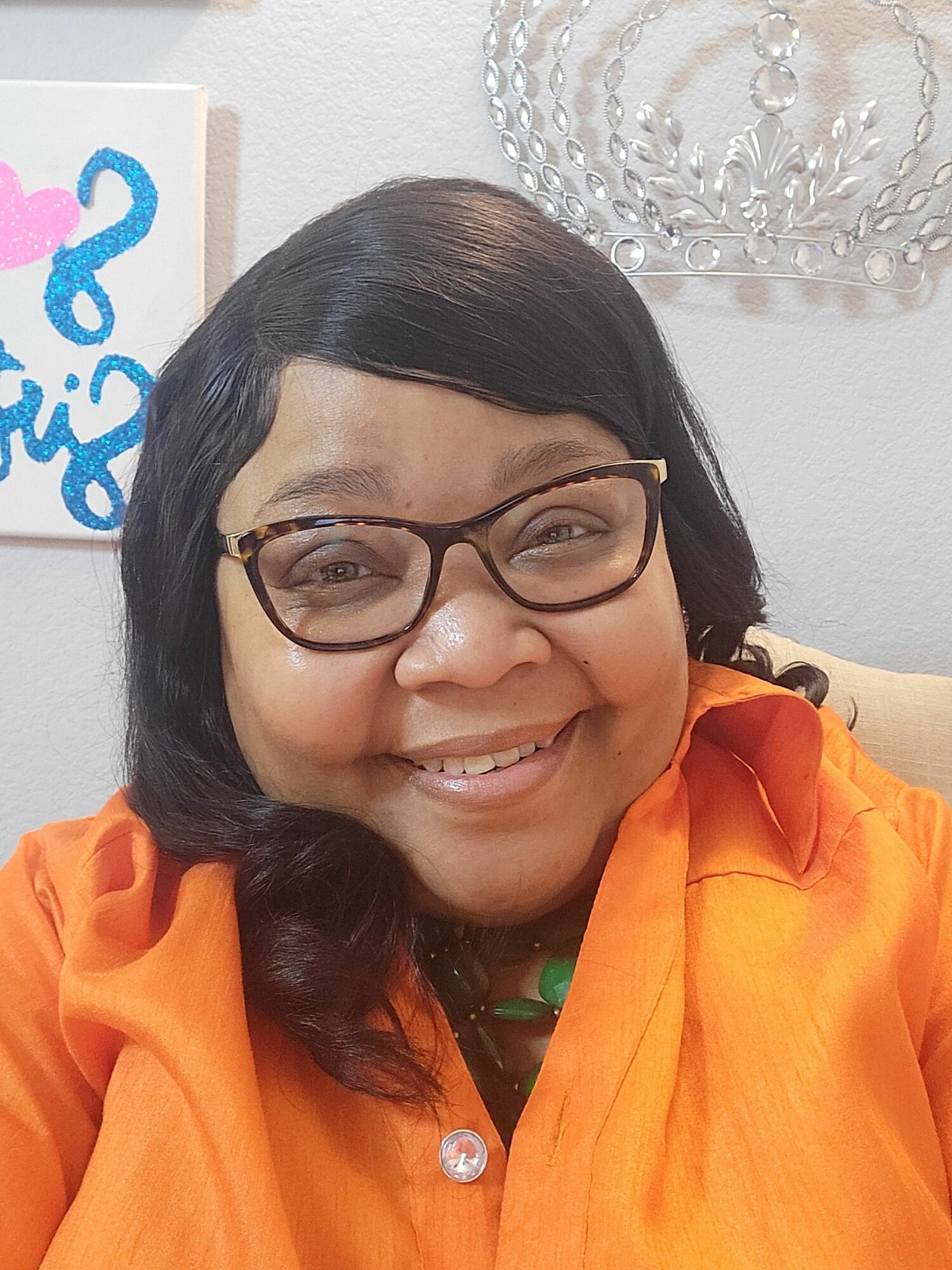 Smiling woman with glasses and dark hair, wearing an orange top, indoors with a decorative wall piece behind her.