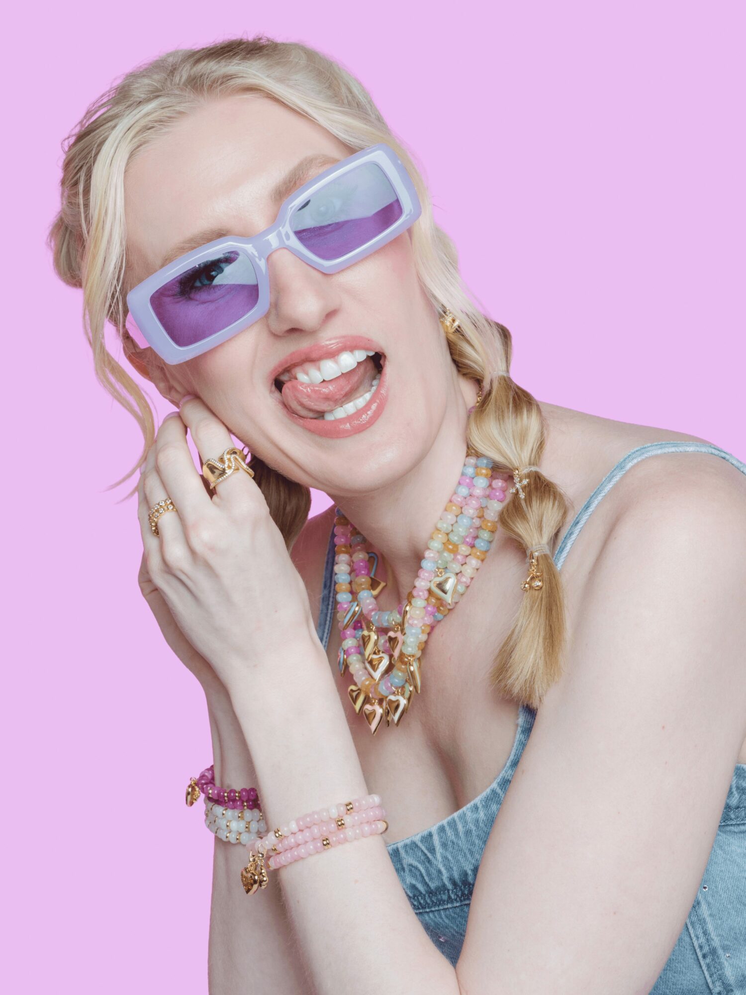 Young woman with blonde hair, wearing purple sunglasses, colorful jewelry, and a denim top, smiling against a pink background.