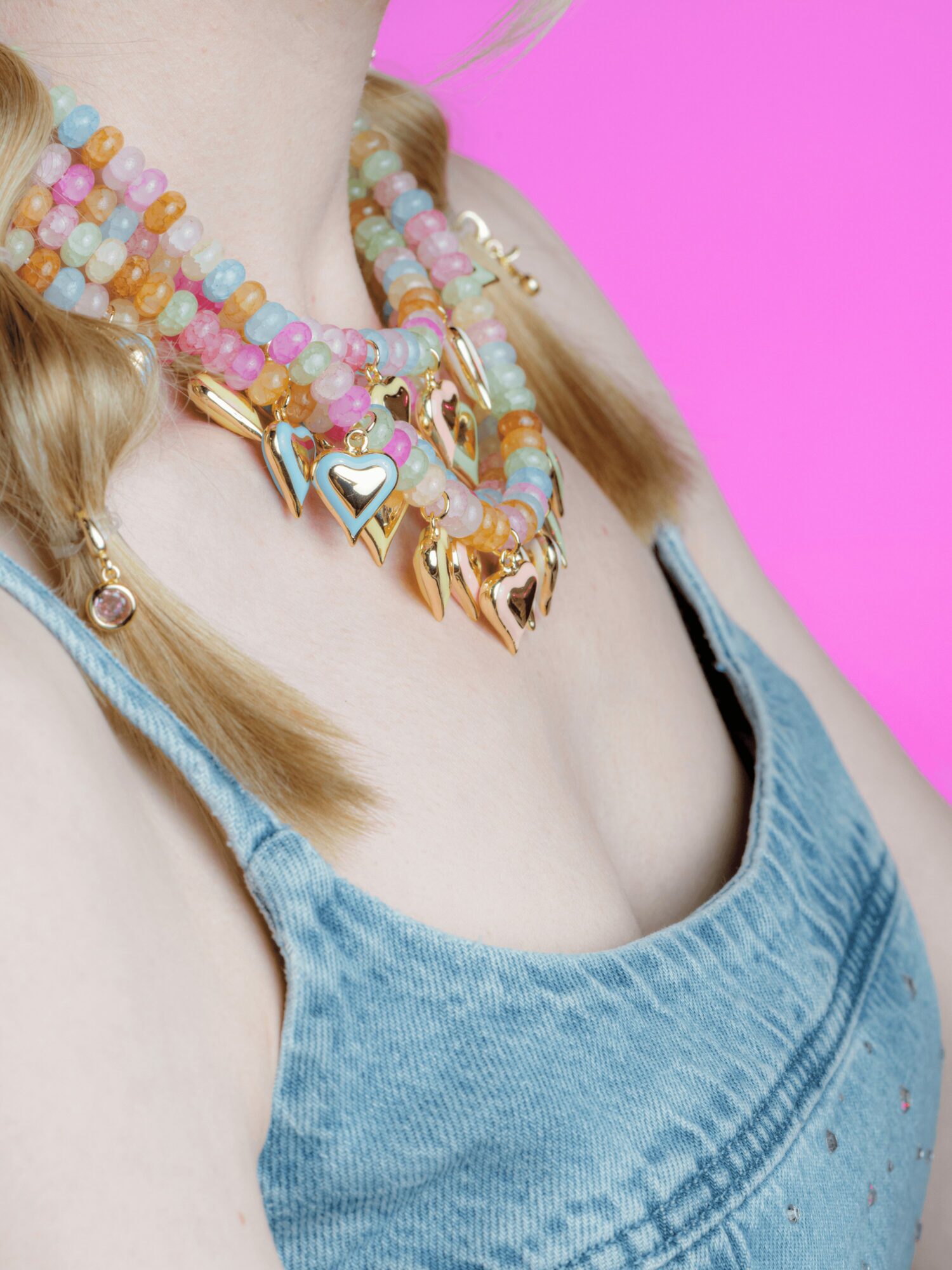Close-up of a person wearing layered colorful beaded necklaces with heart and star charms, against a pink background.