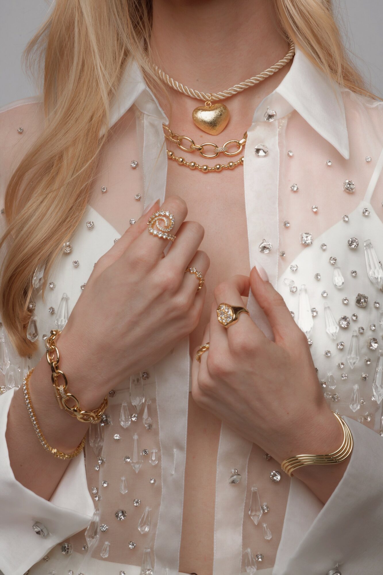 Close-up of a woman wearing layered gold necklaces, rings, and bracelets, with a white shirt adorned with pearls and beads.