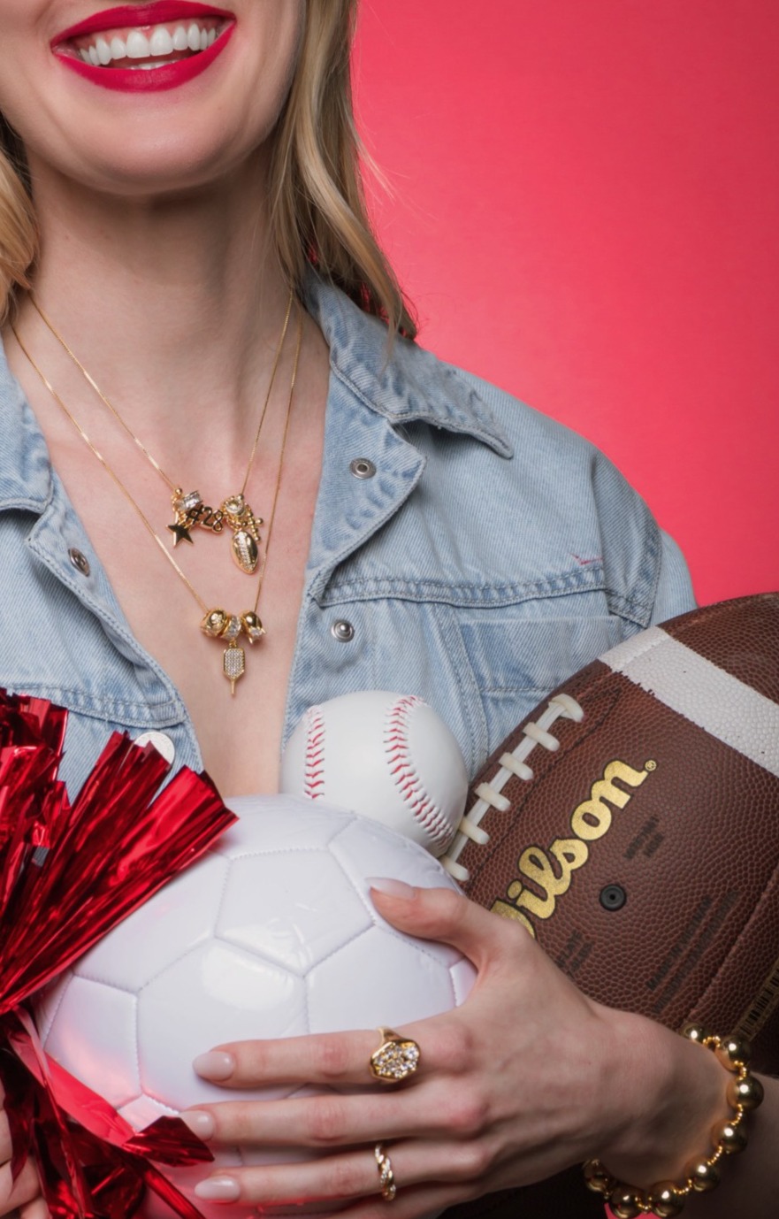 Person holding a baseball, football, and soccer ball, wearing a denim shirt and jewelry, against a pink background.