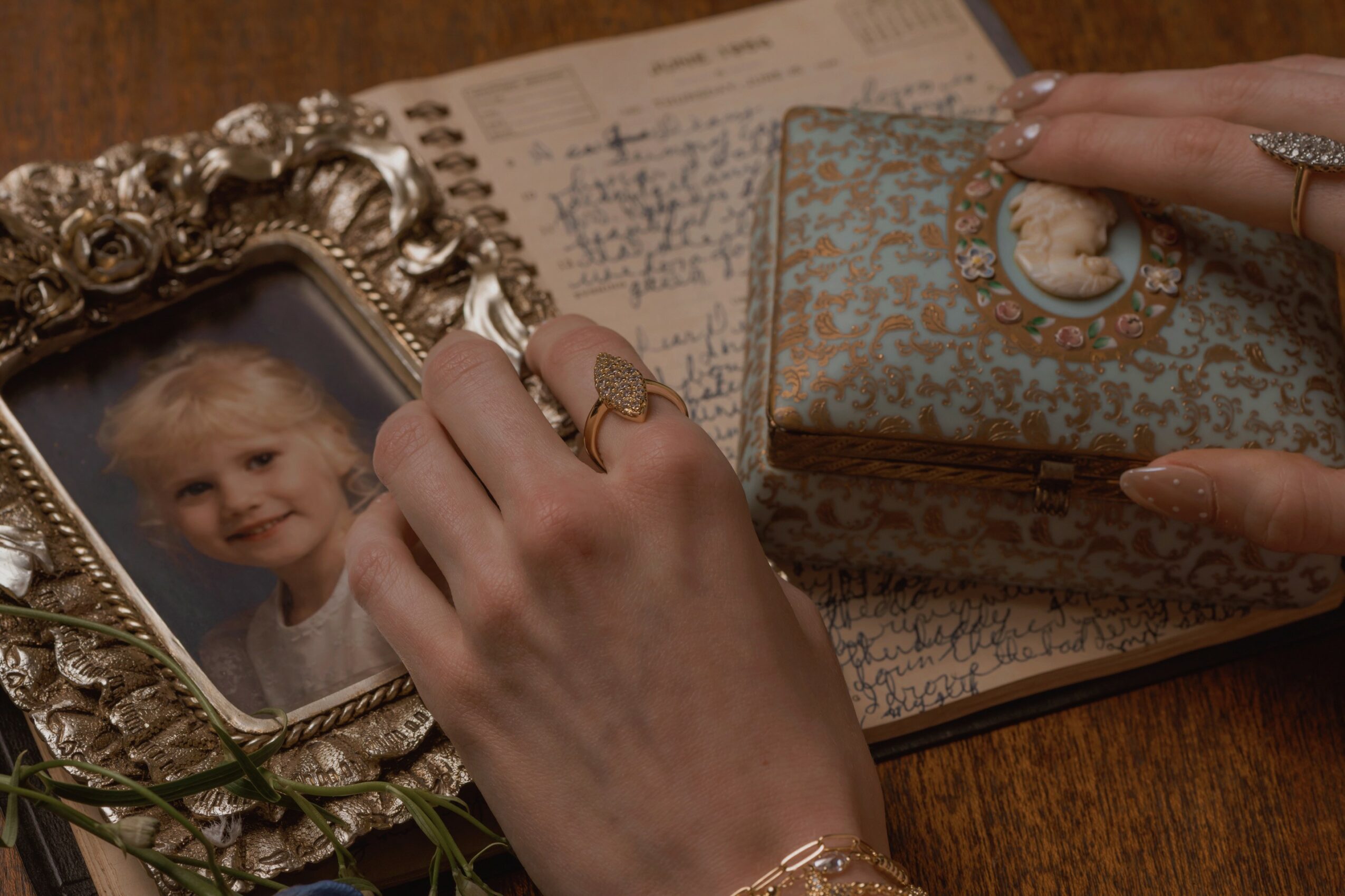 Person's hand with rings holding a photo frame, surrounded by a notebook, a box, and a hand with a ring, on a wooden surface.