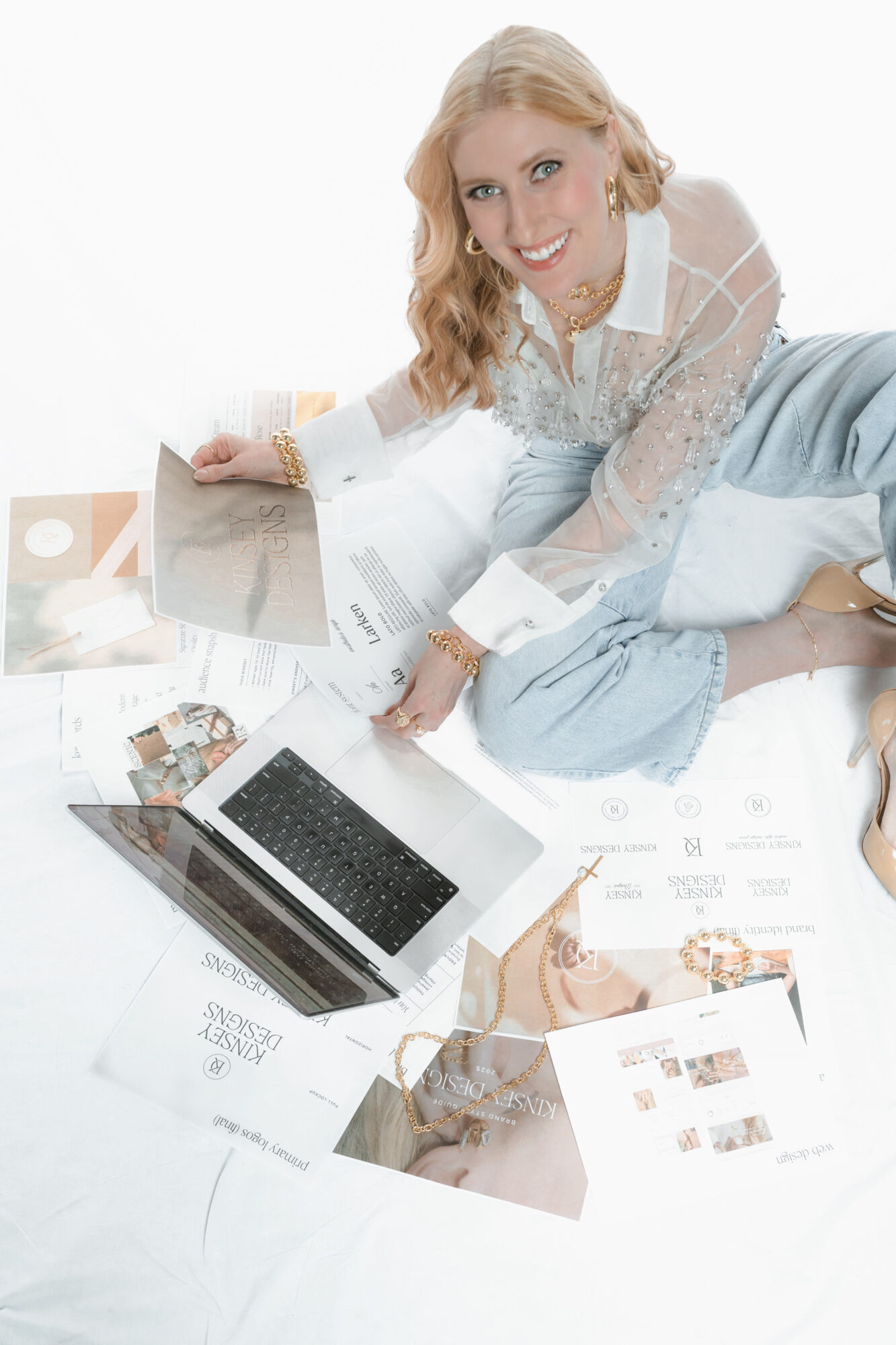 Woman lying on her side on a bed surrounded by papers and a laptop, smiling at the camera.