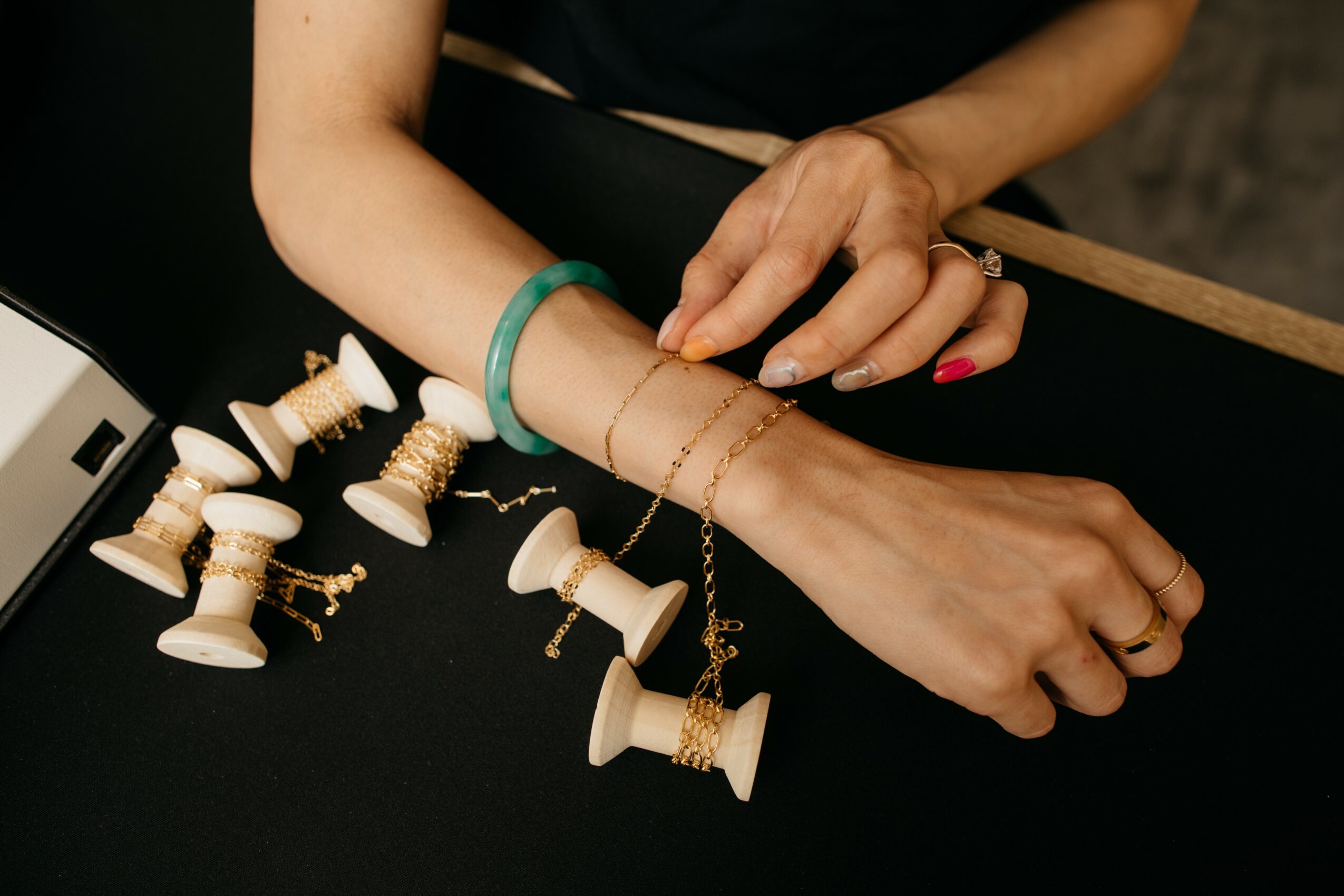 Close-up of a woman's hands with jewelry, jewelry spools, and a black surface, with a blurred background.