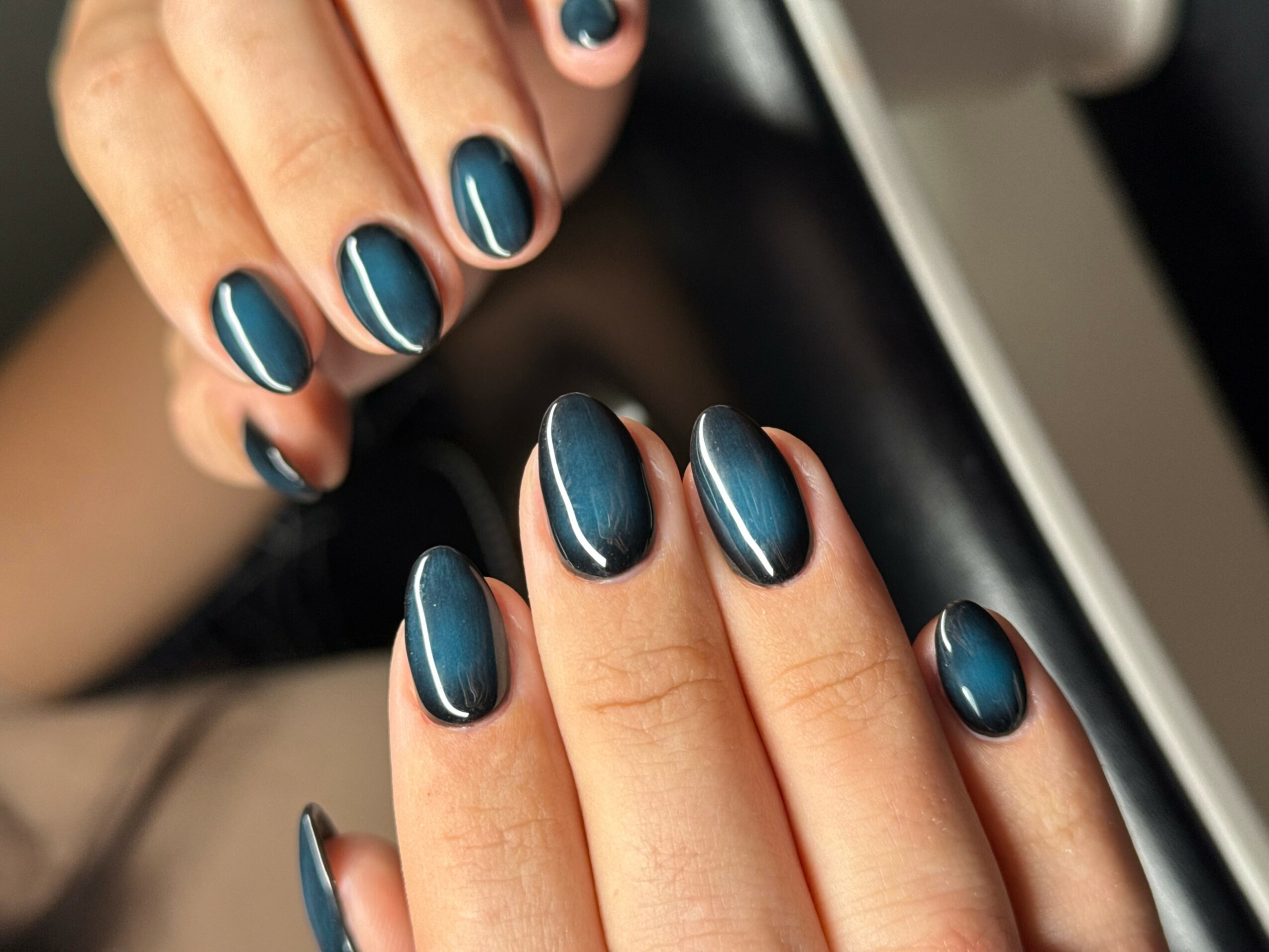 Close-up of hands with dark blue, shiny, oval-shaped nails, resting on a surface.